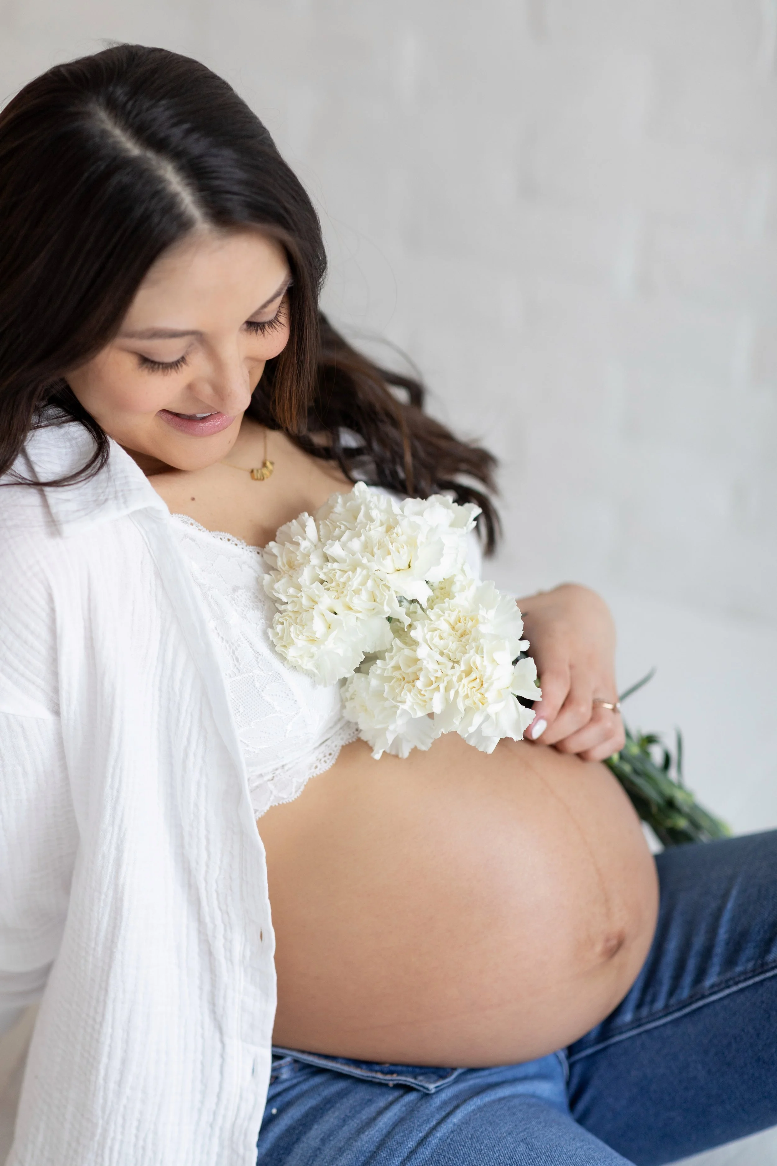 Pregnant woman holding white flowers, sitting in a white top and blue jeans with a visible belly.