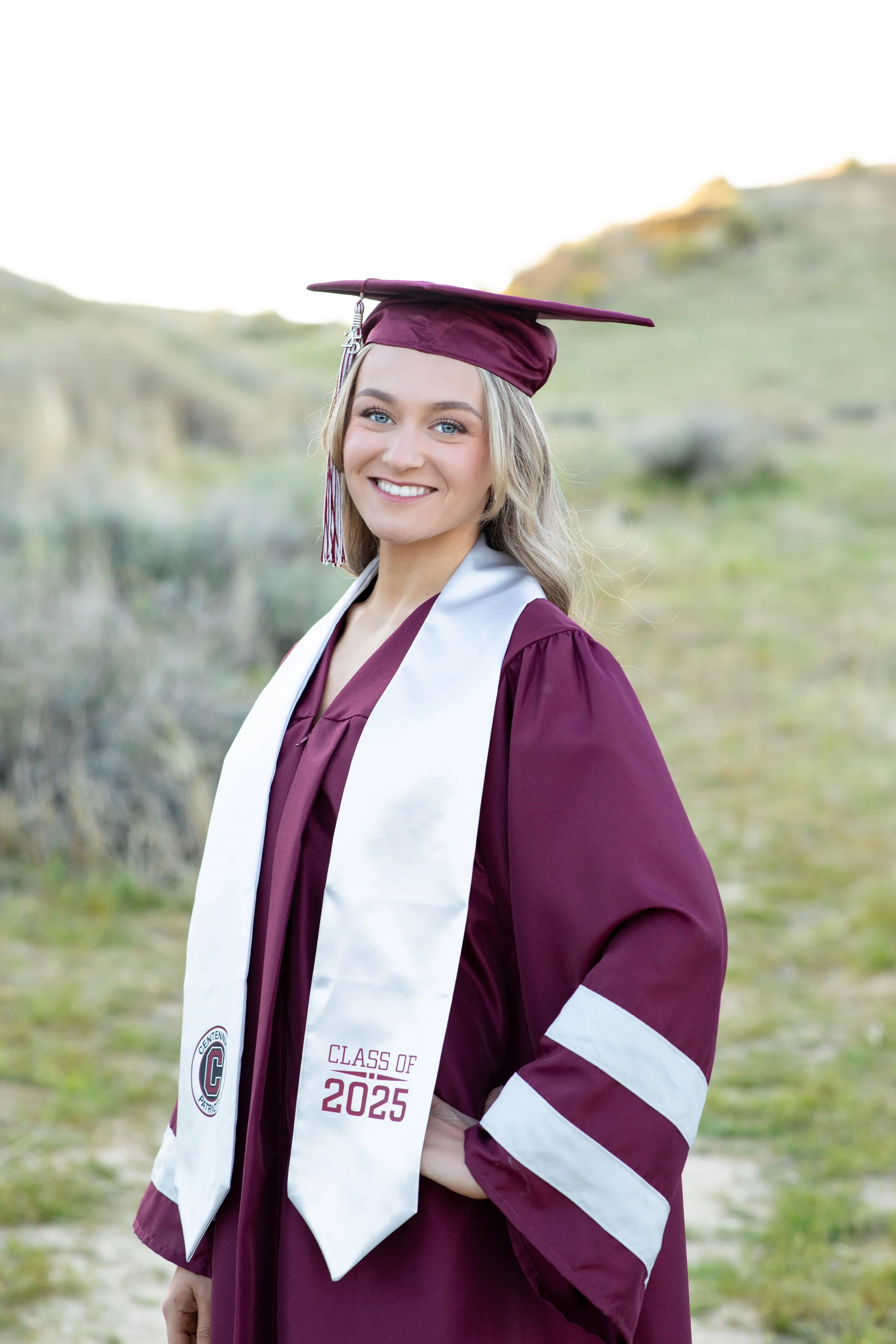 A young woman in a maroon graduation gown with a white sash that says "Class of 2025" stands outdoors on a grassy path, smiling at the camera, wearing a matching maroon cap with a tassel.