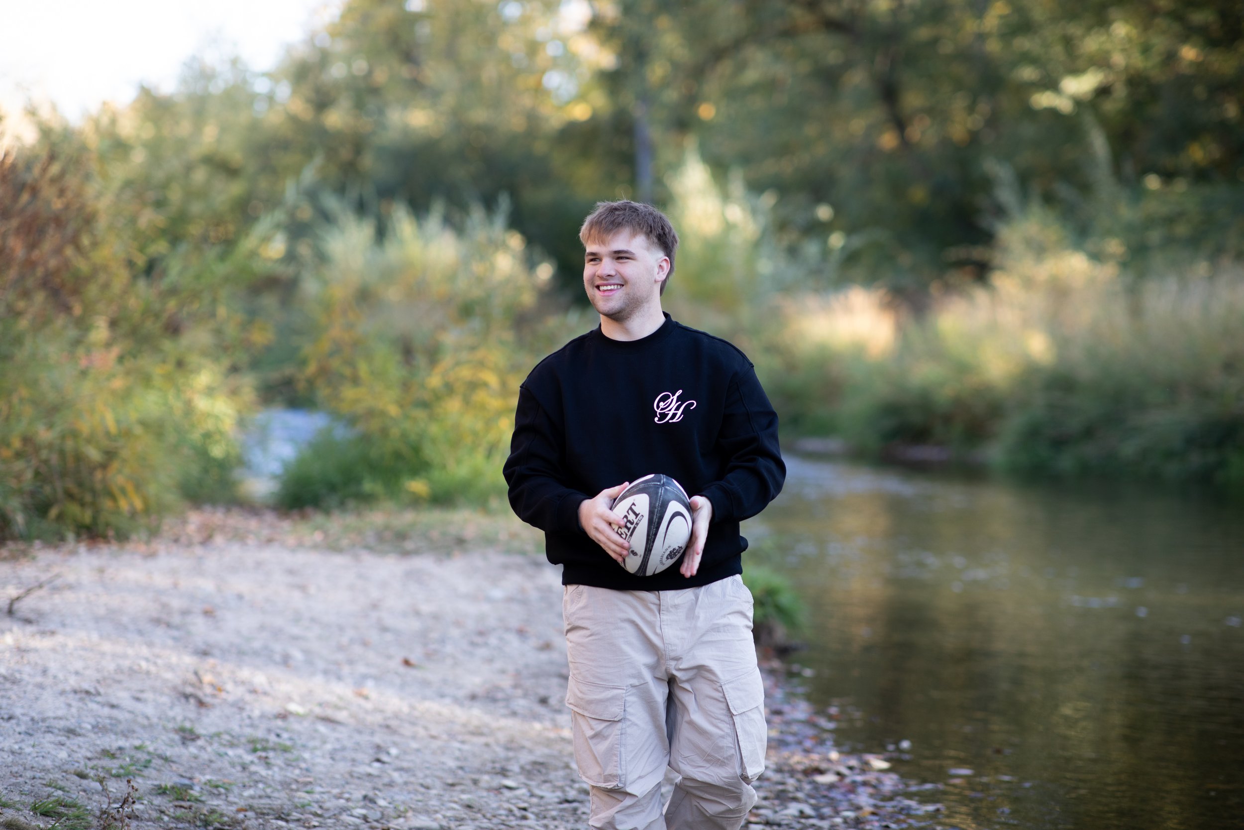 A young man holding a rugby ball, walking along a riverbank with trees and foliage in the background on a sunny day.