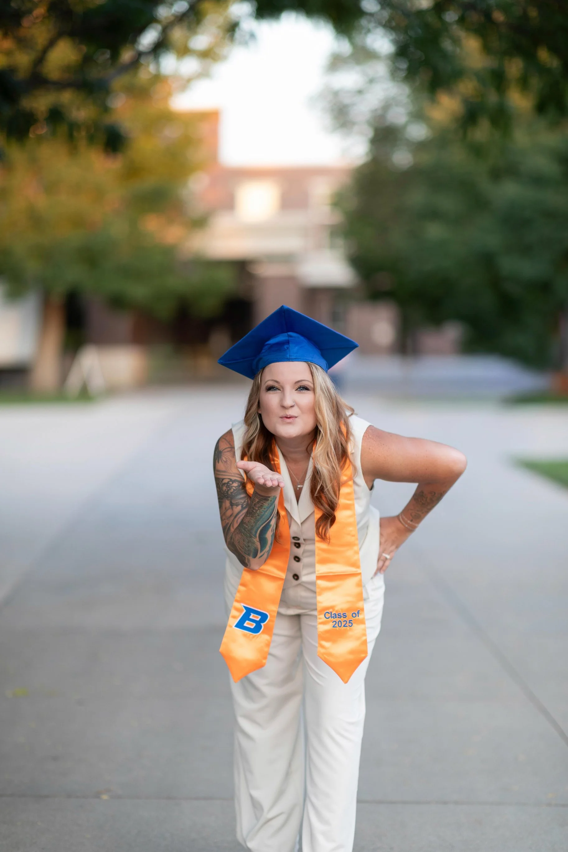 Young woman in a white outfit with tattoos, wearing a blue graduation cap and orange sash that says 'Class of 2025,' blowing a kiss.