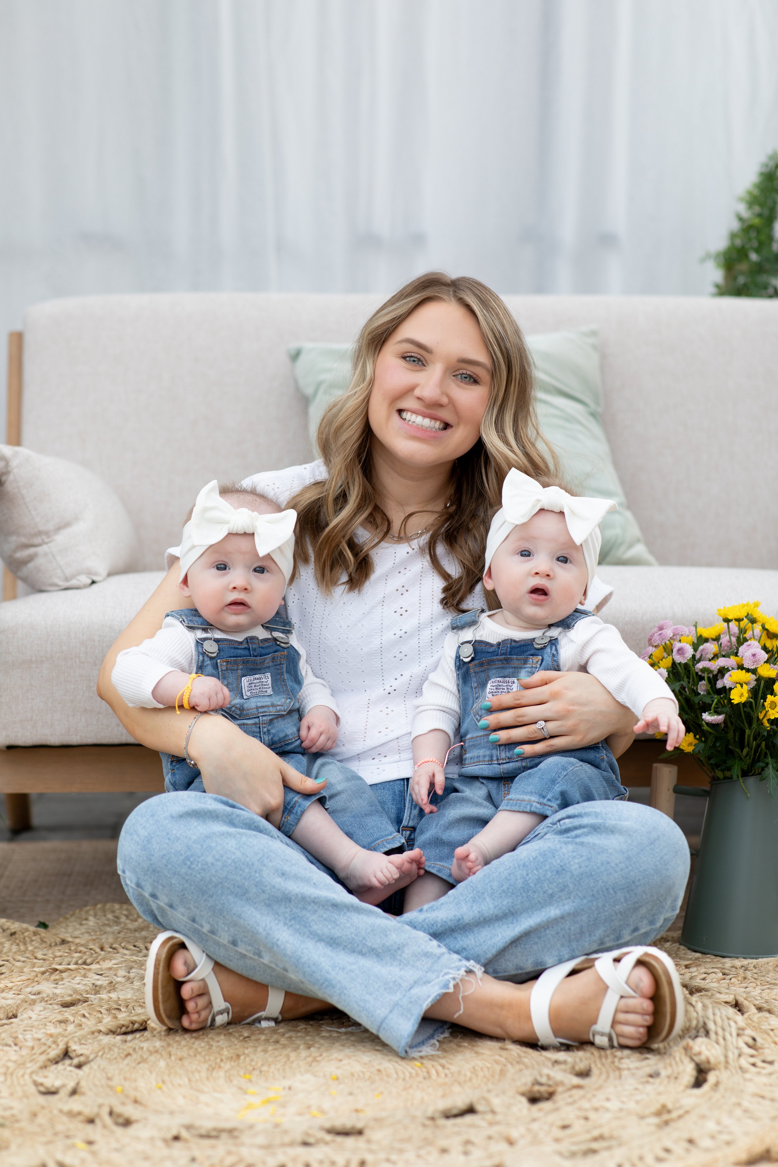 A woman sitting cross-legged on the floor, smiling, holding two babies dressed in denim overalls and white tops, each with a white bow headband, in a cozy living room with a beige sofa, cushions, a potted plant, and a vase of flowers nearby.