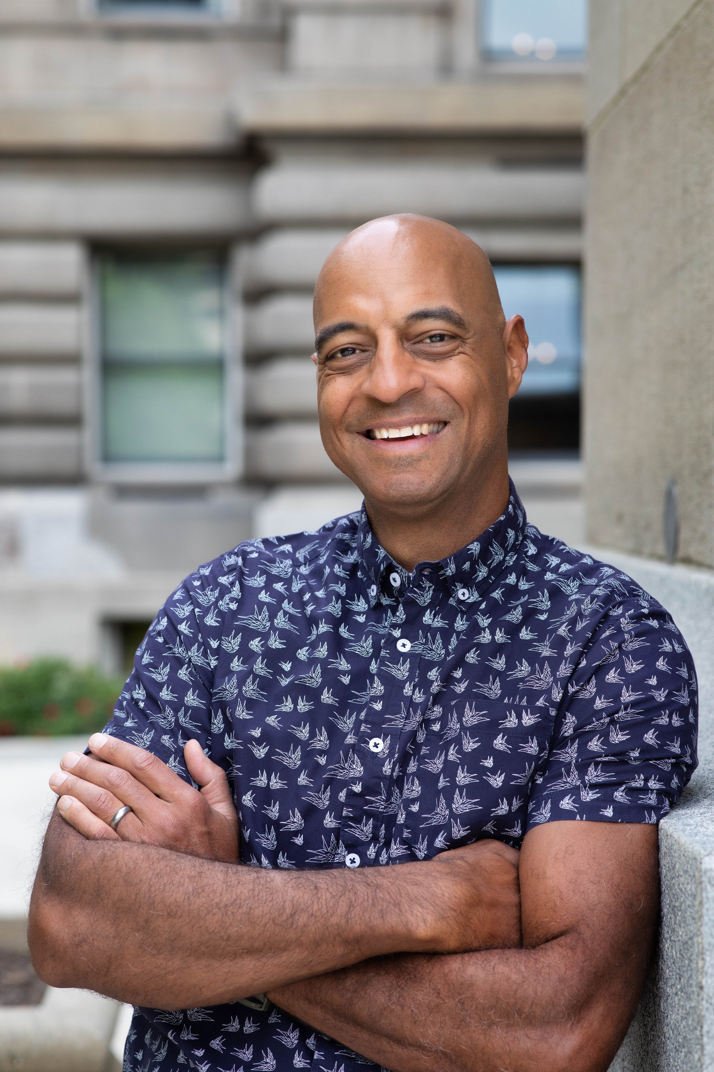A smiling man with crossed arms standing outdoors in front of a building with modern architecture, wearing a navy blue shirt with a white leaf pattern.