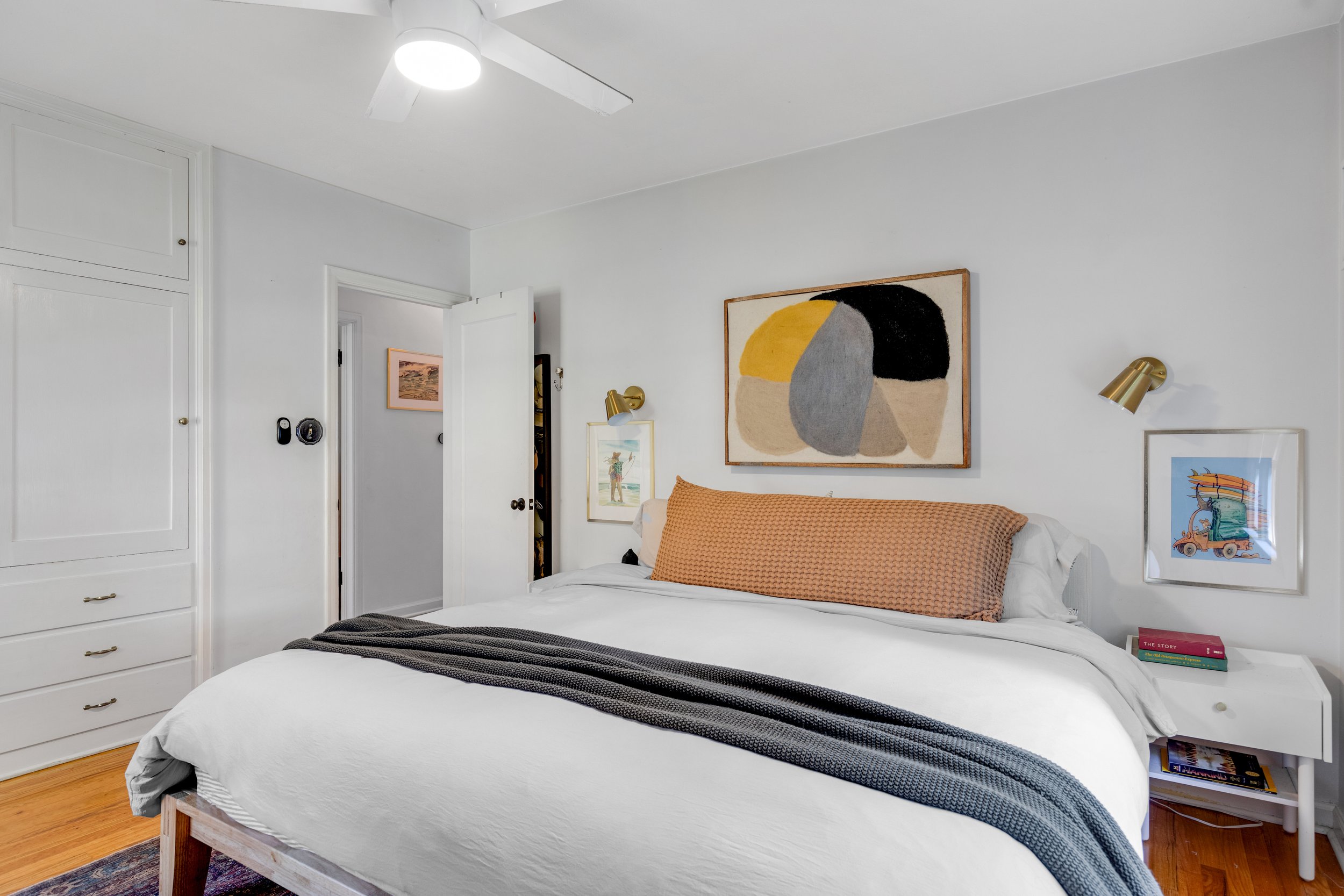 A neatly made bed with white bedding, a large brown textured pillow, and a dark gray blanket. Above the bed are three framed pictures and two gold wall-mounted reading lights. To the right is a white nightstand with a couple of books and a colorful a