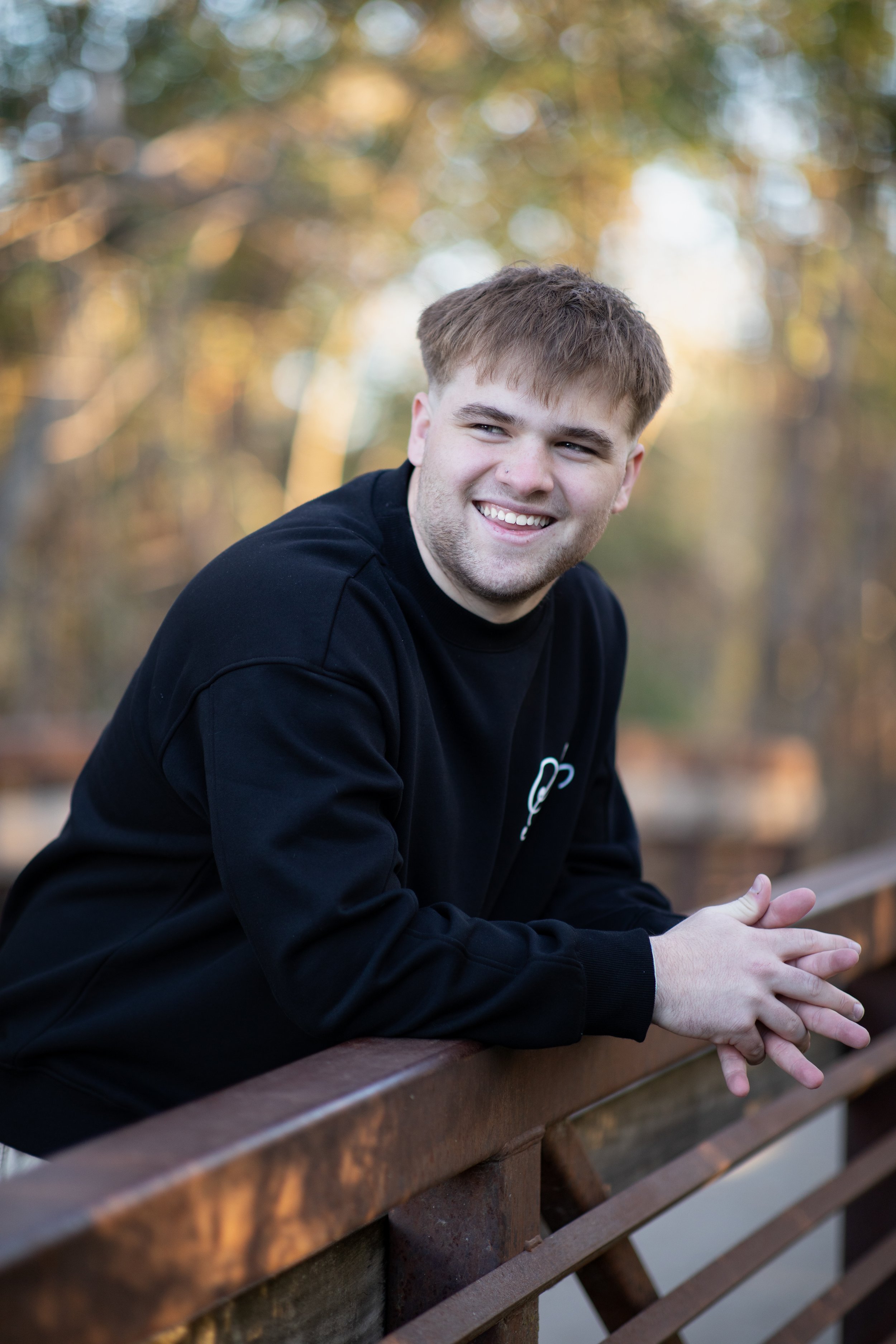 A young man with short brown hair and a beard smiling while leaning on a wooden railing outdoors during autumn.