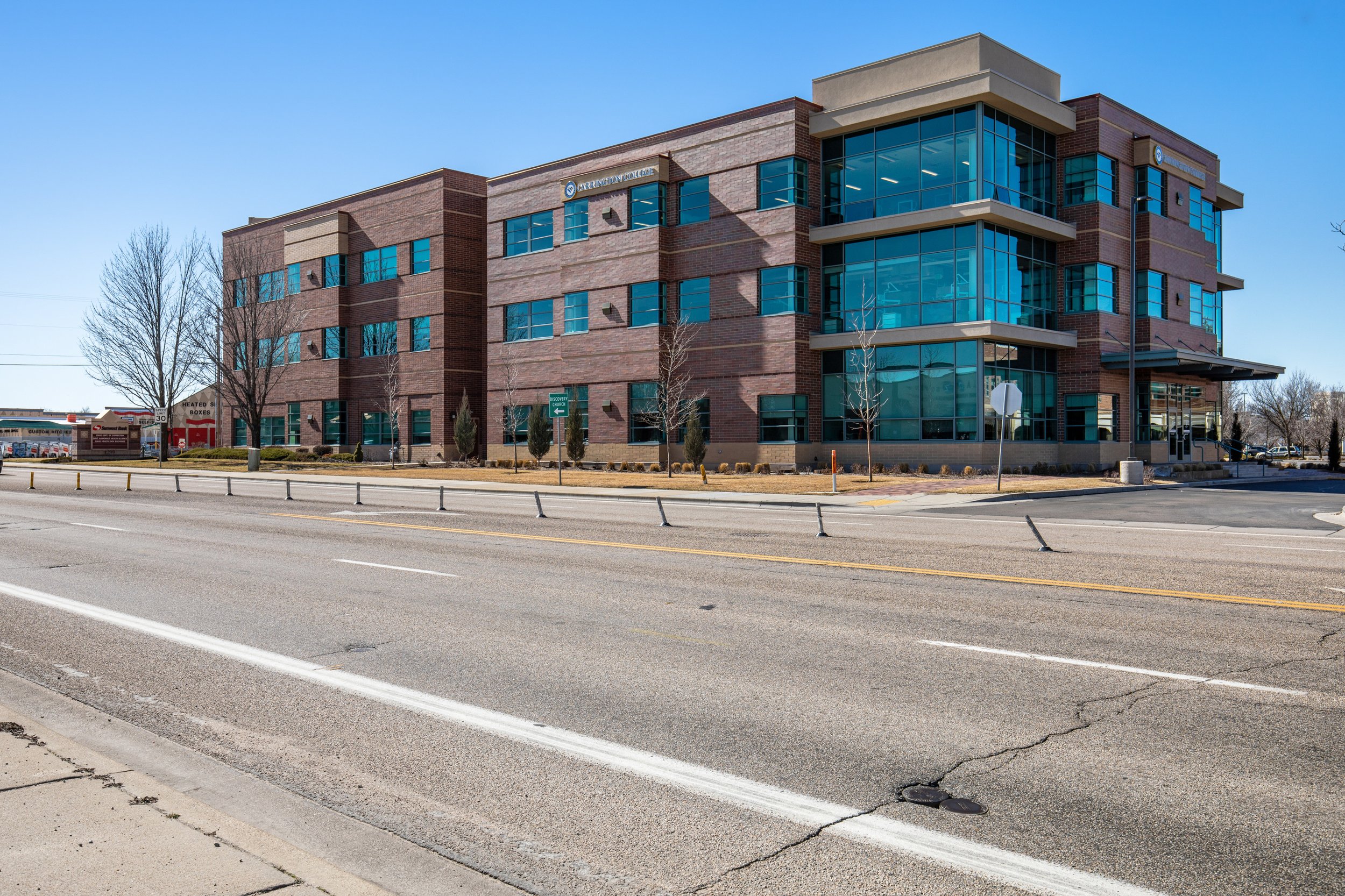 A large, modern brick building with multiple floors and large glass windows, located next to a wide street with traffic barriers and a crosswalk.