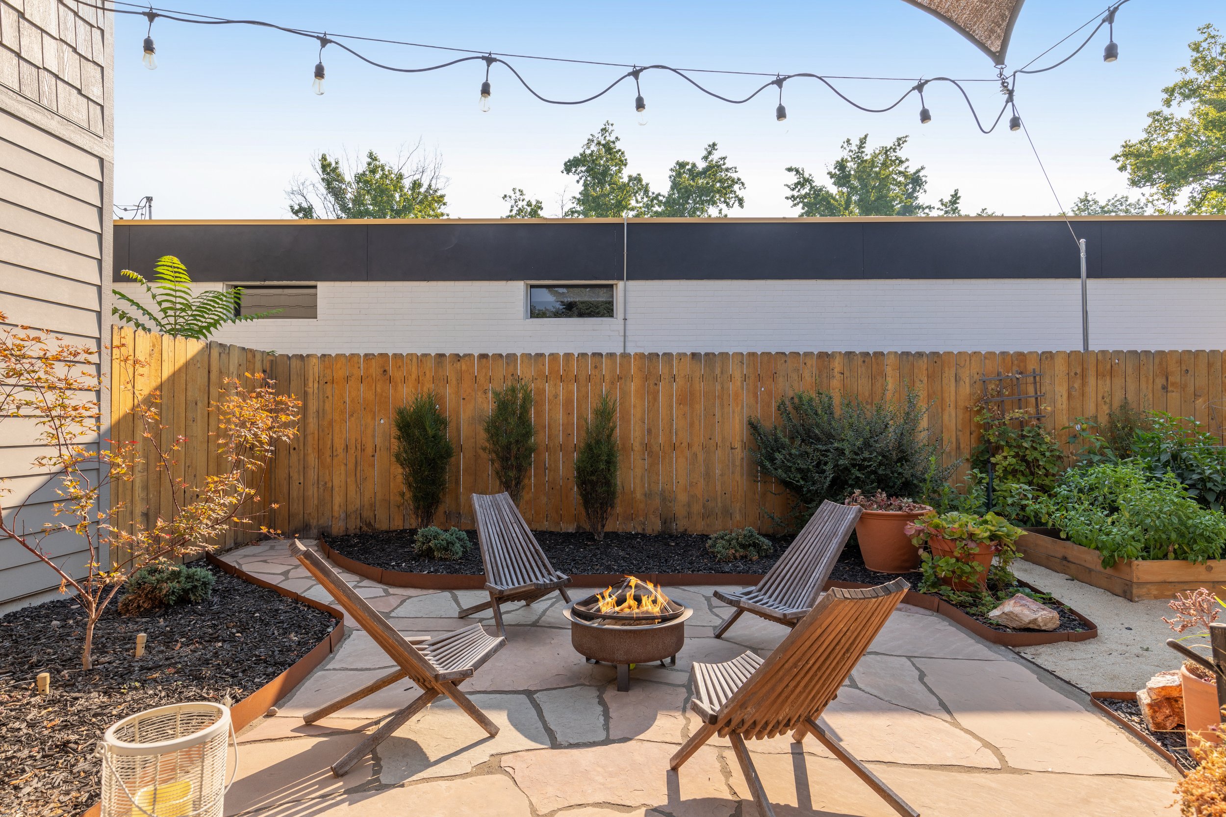 Backyard patio with four wooden chairs arranged around a small fire pit, bordered by a wooden fence, potted plants, and string lights overhead.