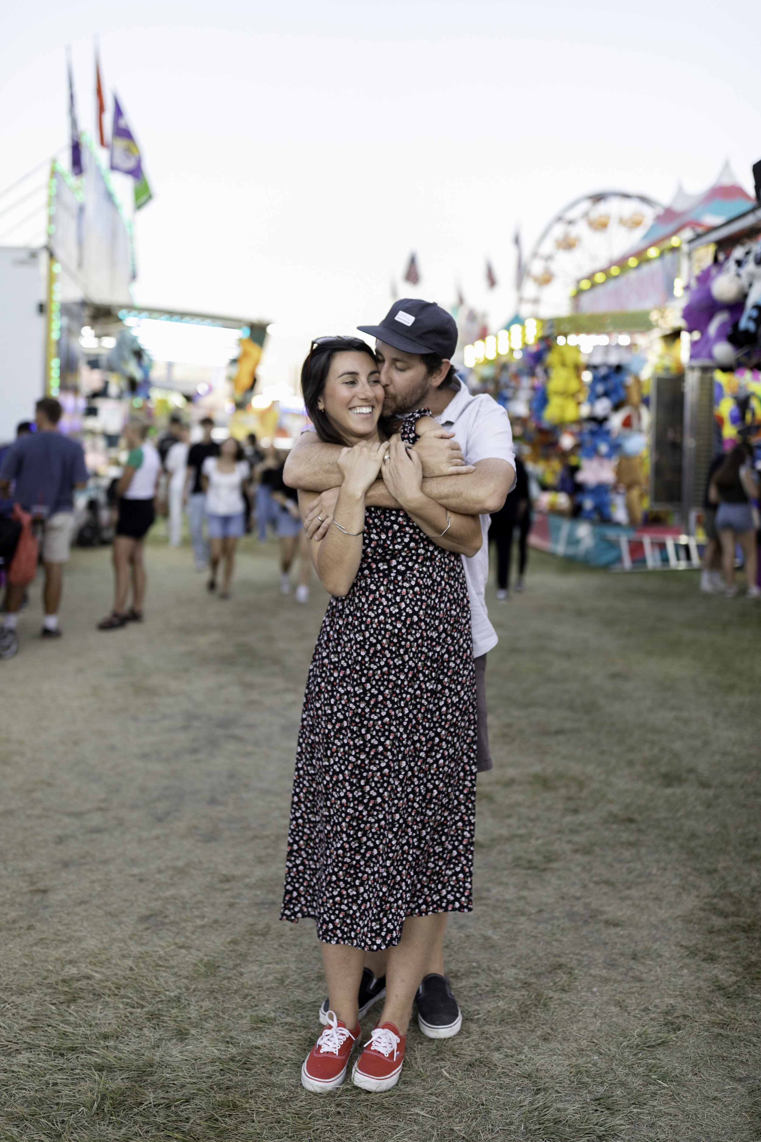 A couple hugging and smiling at a funfair or carnival, with colorful rides and game booths in the background, during daylight.