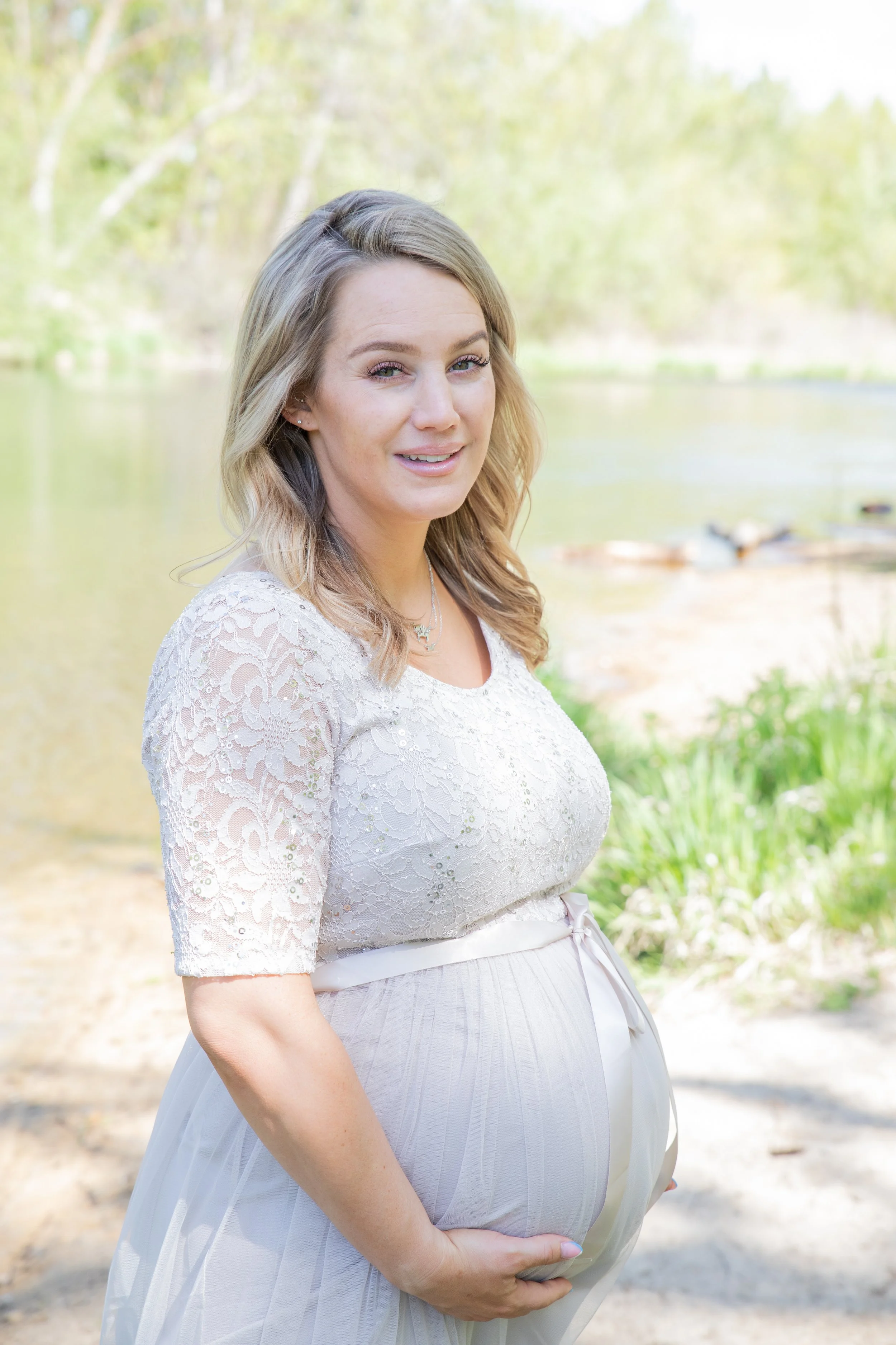 A pregnant woman standing outdoors near a body of water, holding her belly with one hand, wearing a white lace top and light-colored skirt, with trees and water in the background.