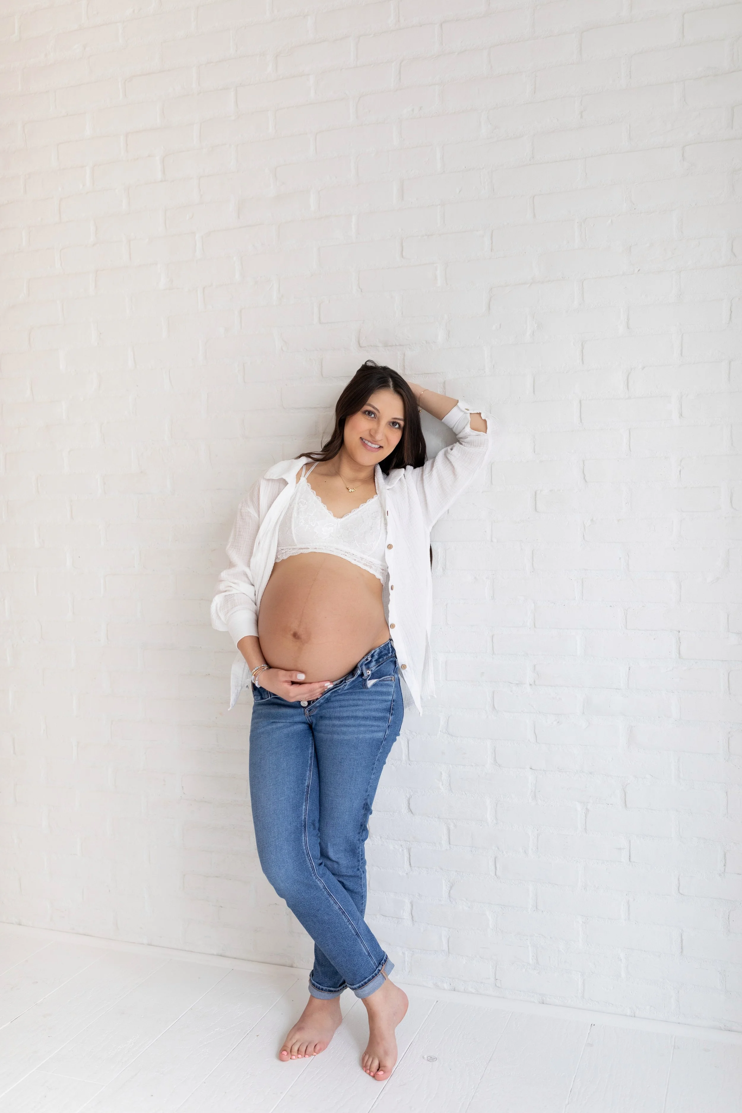 Pregnant woman in a white bra and unbuttoned white shirt, standing barefoot against a white brick wall, smiling, with one hand on her belly and the other resting behind her head.