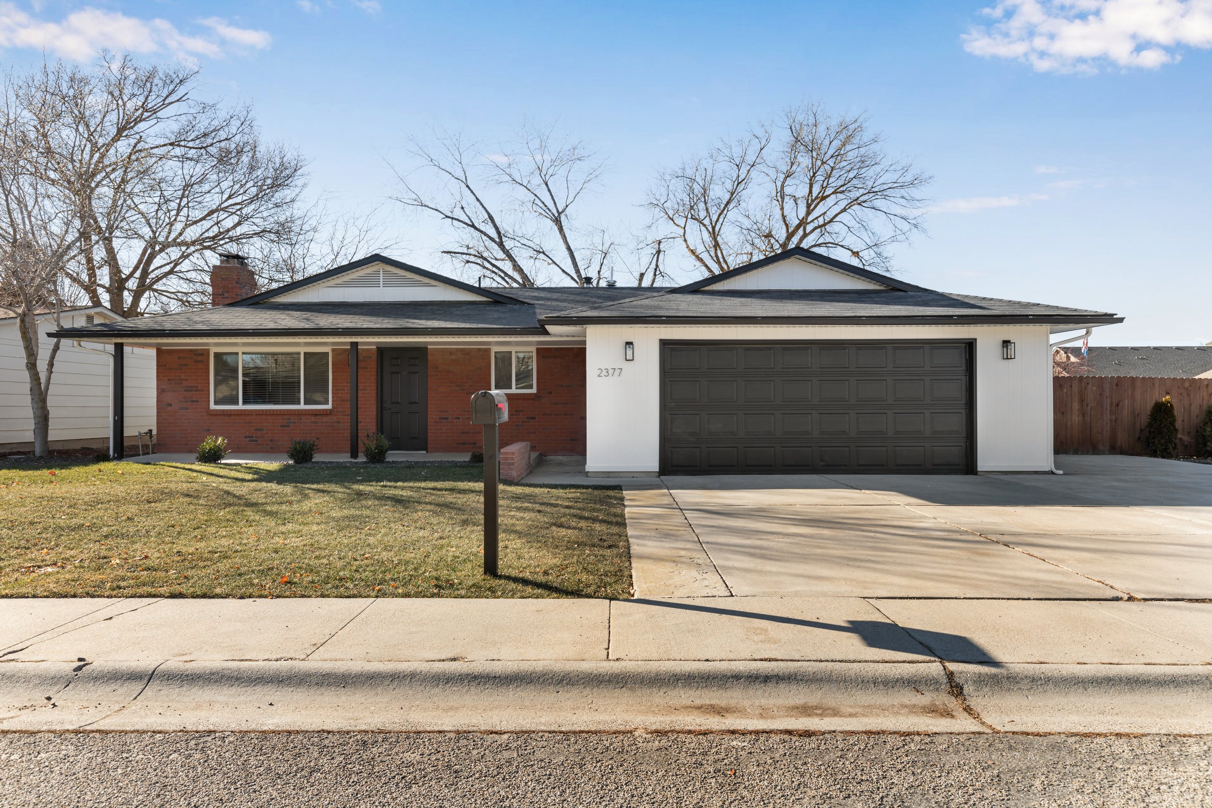 Single-story house with a red brick facade and white siding, black garage door, front door, and trim, small front lawn with a mailbox, and leafless trees in the background under a clear blue sky.