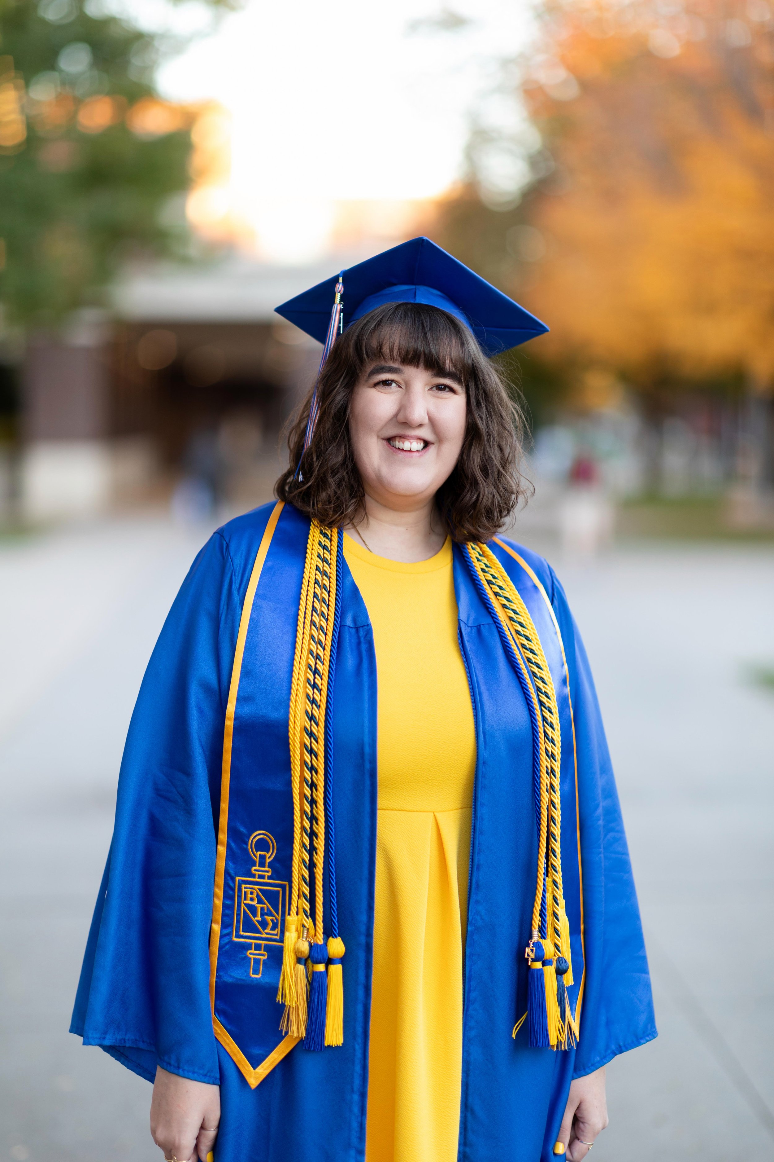 A young woman in a blue graduation gown and cap, with yellow accents and cords, smiling outdoors during sunset with autumn-colored trees in the background.