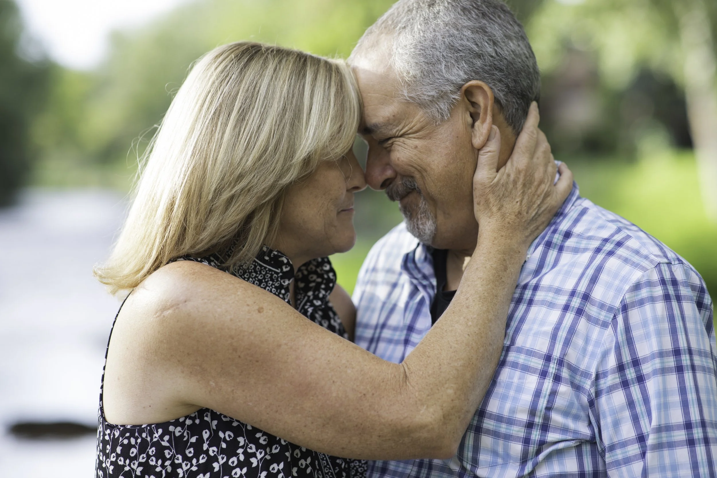 An older couple with gray hair and light skin touching foreheads and embracing outdoors with blurred green trees in the background.