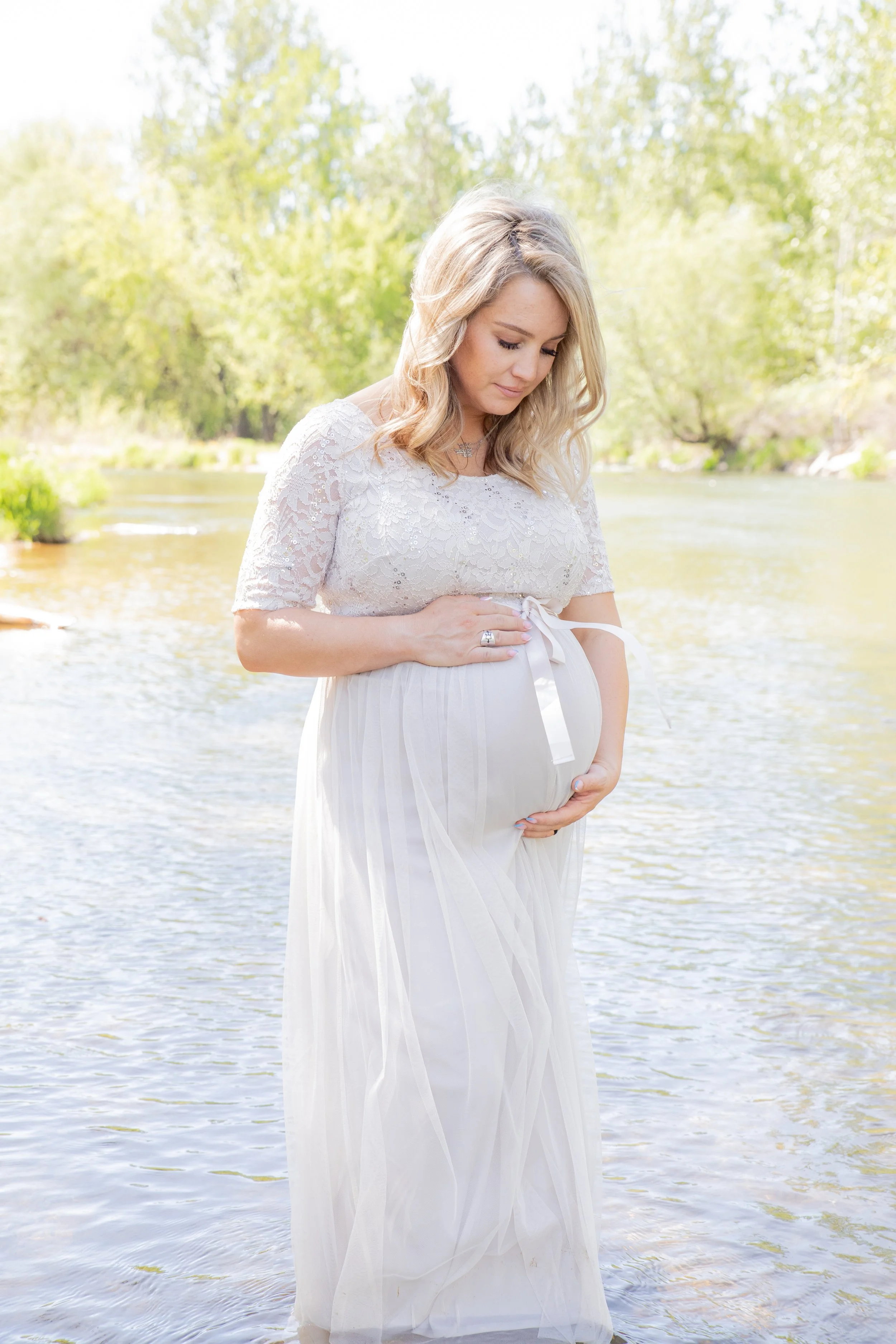 A pregnant woman wearing a white lace dress standing in a river, gently touching her belly with both hands, surrounded by greenery and trees.