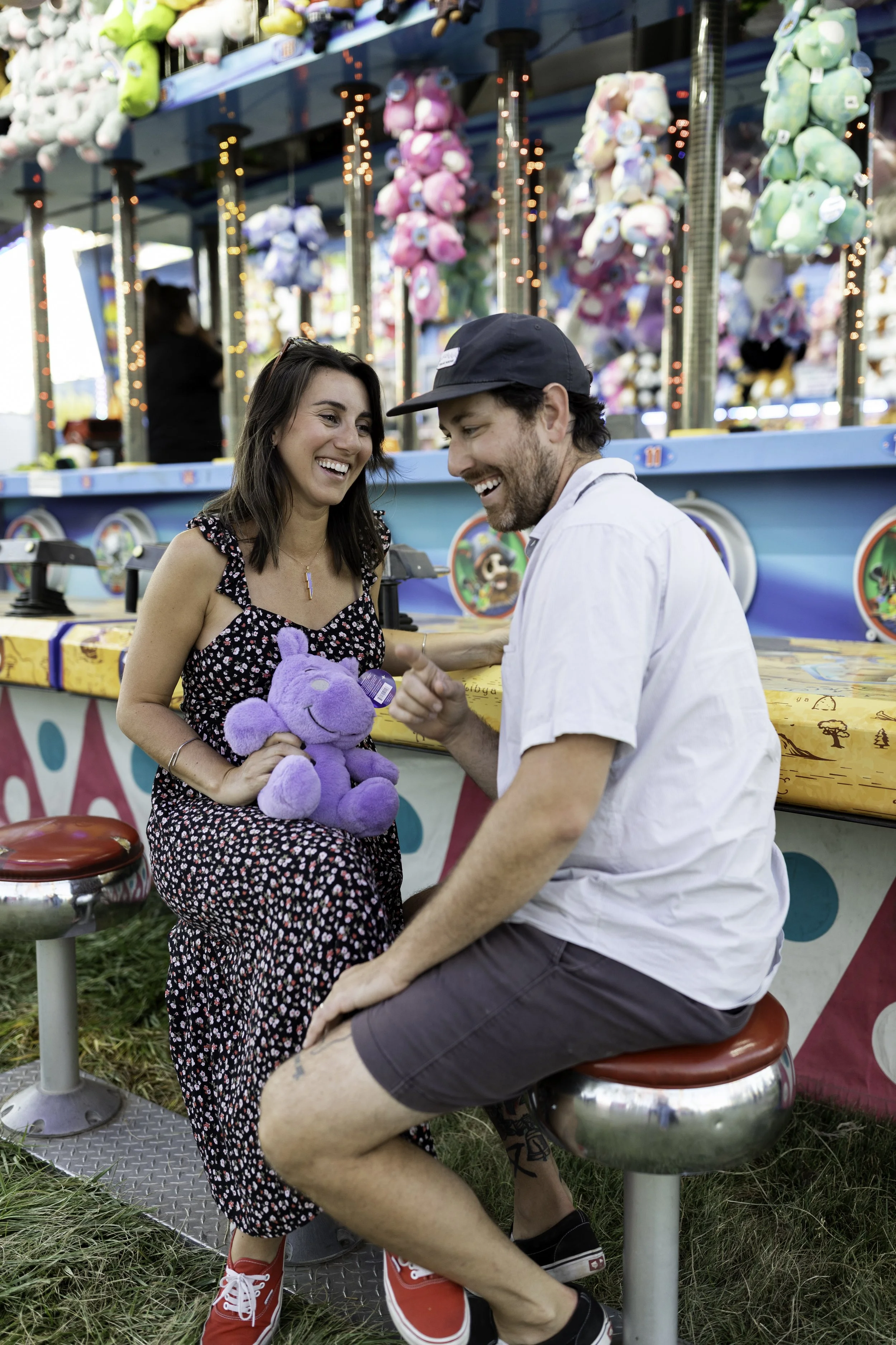 A woman and a man are laughing together at a carnival game booth. The woman is holding a purple stuffed animal, and there are colorful plush toys hanging behind them.