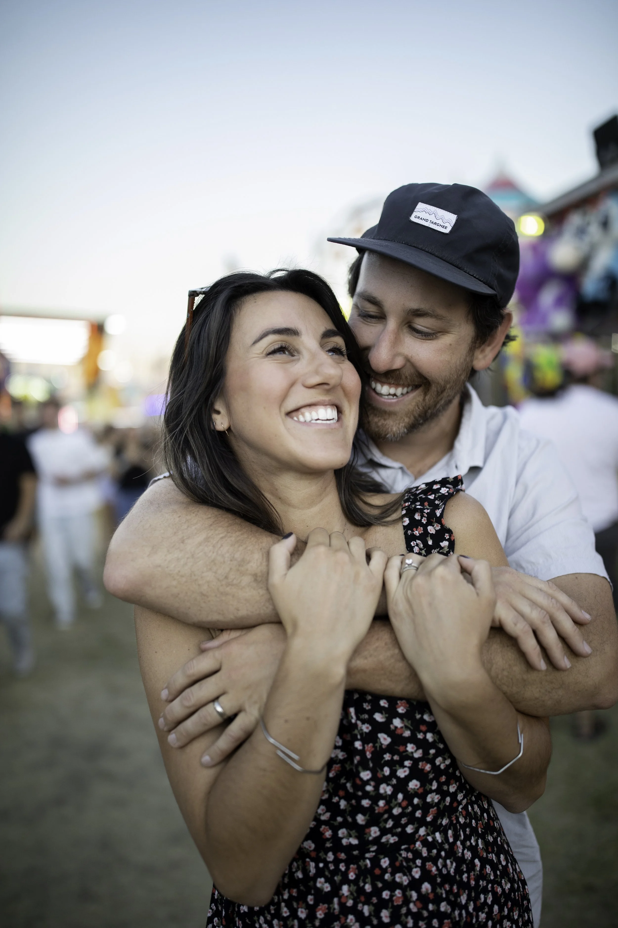 A happy couple embraces at an outdoor festival or fair, with colorful booths and blurred crowd in the background during daylight.