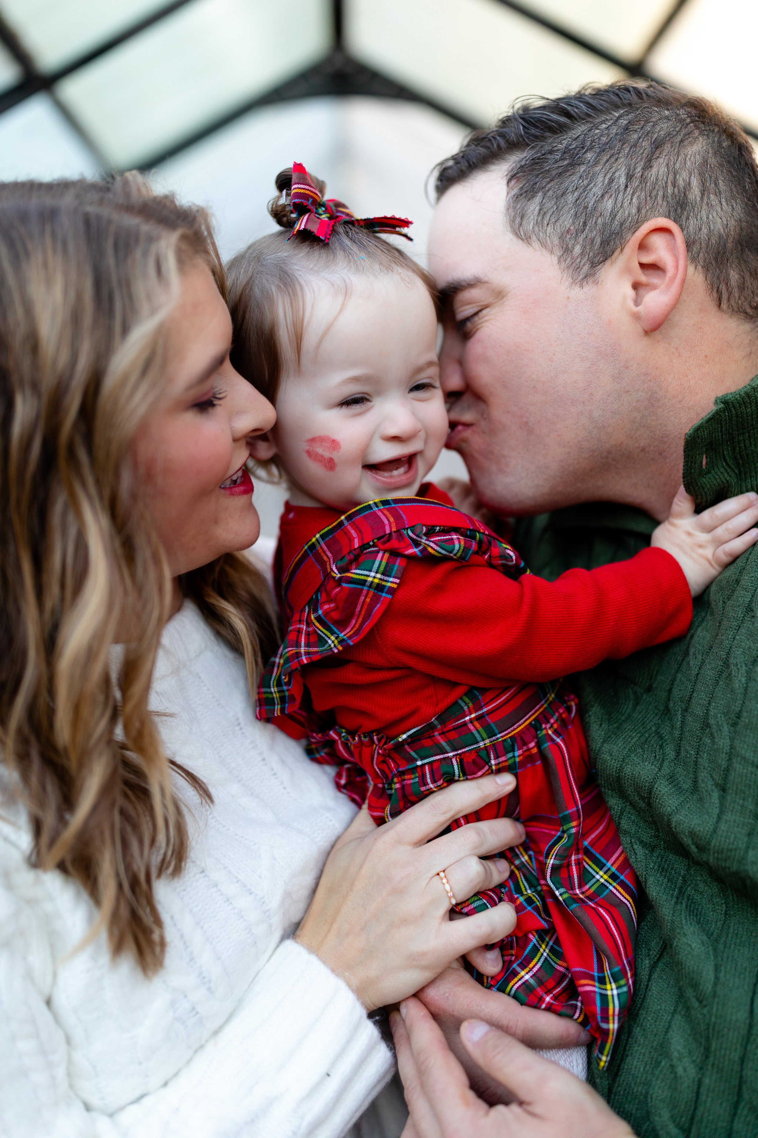 A family of four, including a baby girl with a bow in her hair and lipstick kiss on her cheek, gathered in a close embrace, smiling and celebrating together during the holiday season.