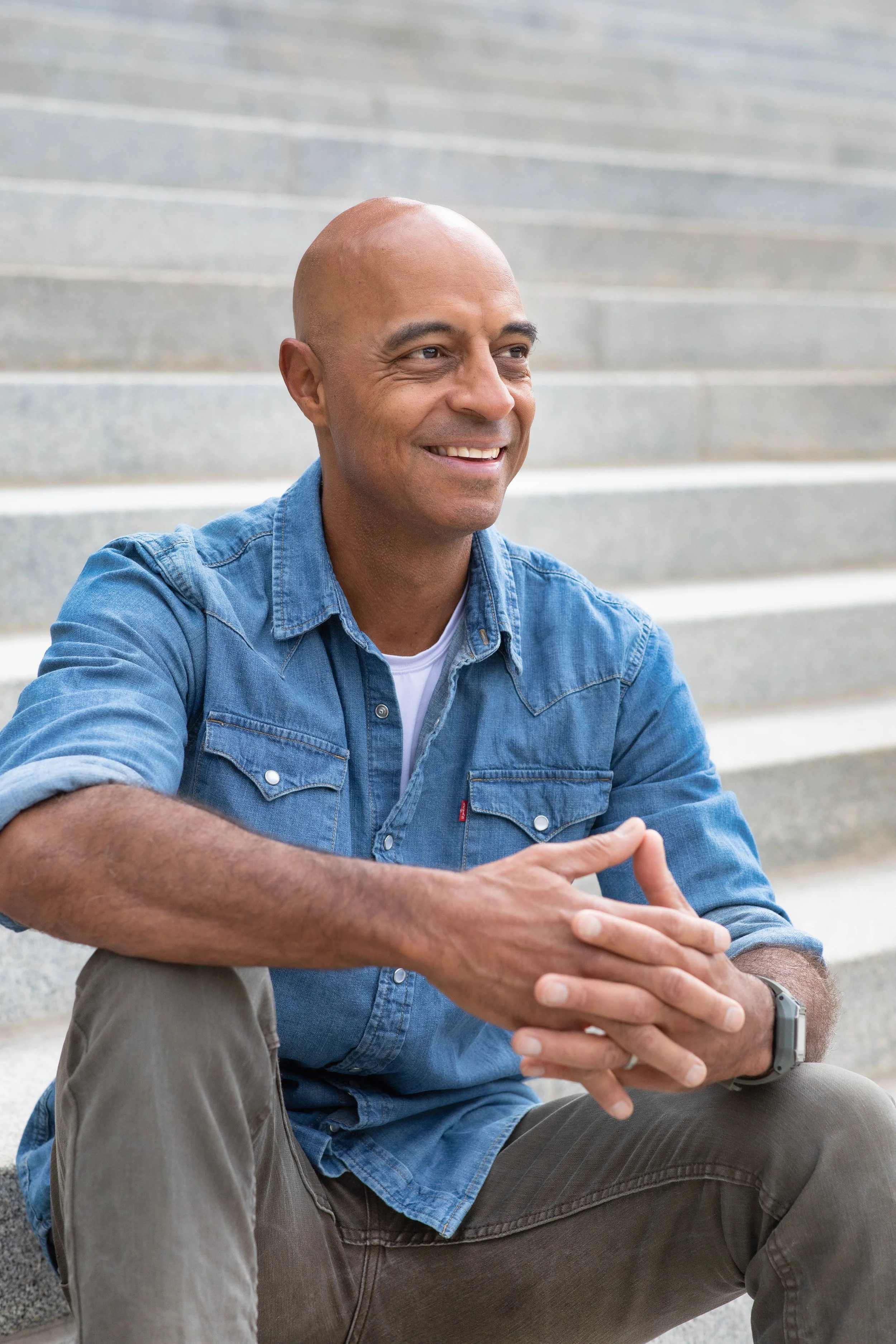 A smiling, bald man in a blue denim shirt sitting on stone steps, looking to the side, with a smartwatch on his wrist.