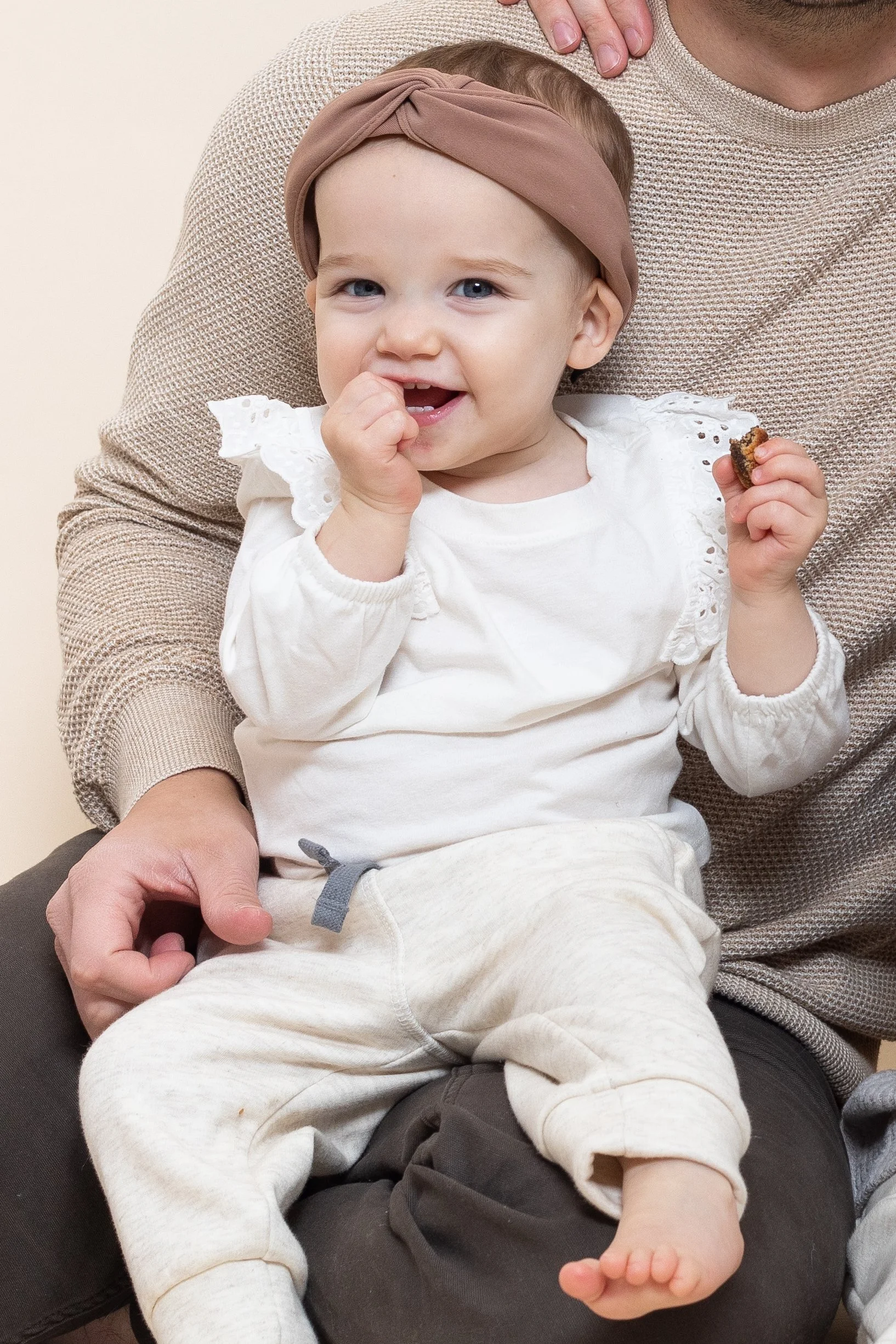 A smiling toddler girl with a brown headband sitting on a man's lap, holding a cookie and wearing a white long-sleeve top with eyelet details on the shoulders and light-colored sweatpants.
