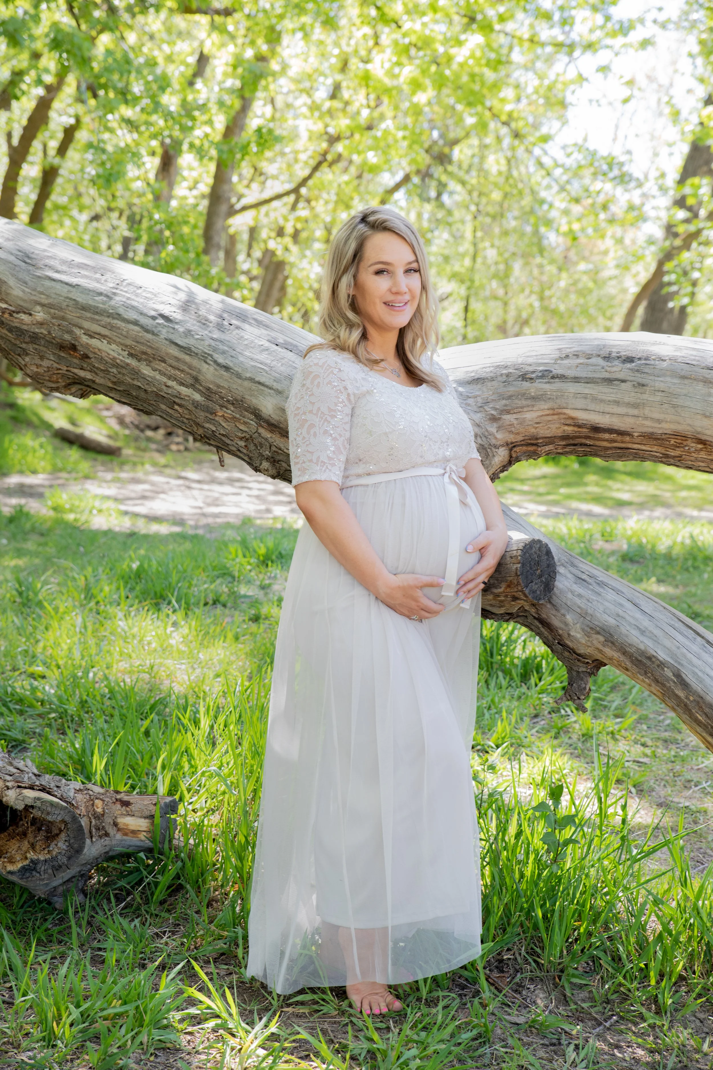 Pregnant woman standing outdoors in a green park, holding her baby bump, next to a fallen tree.