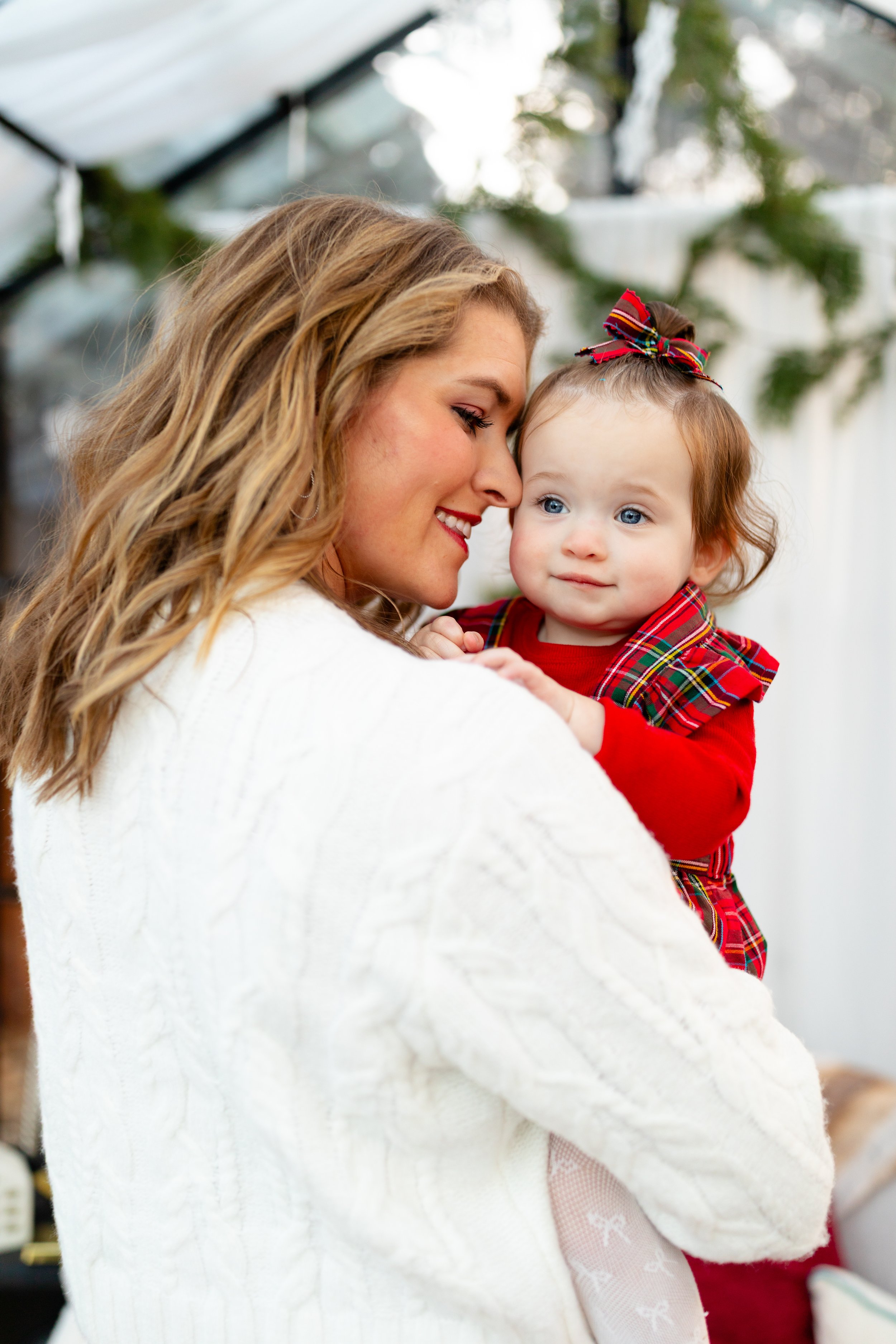 A woman holding a young girl with blue eyes, both smiling, in a bright indoor setting. The girl is dressed in red plaid with a matching bow in her hair.