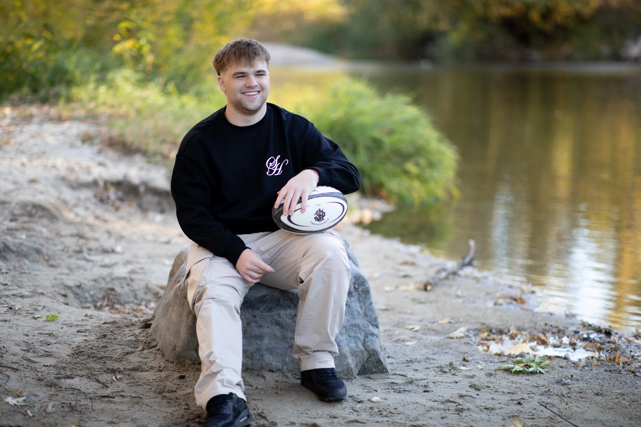 A young man sitting on a large rock by a river, holding a rugby ball, smiling, in an outdoor setting with trees and water.