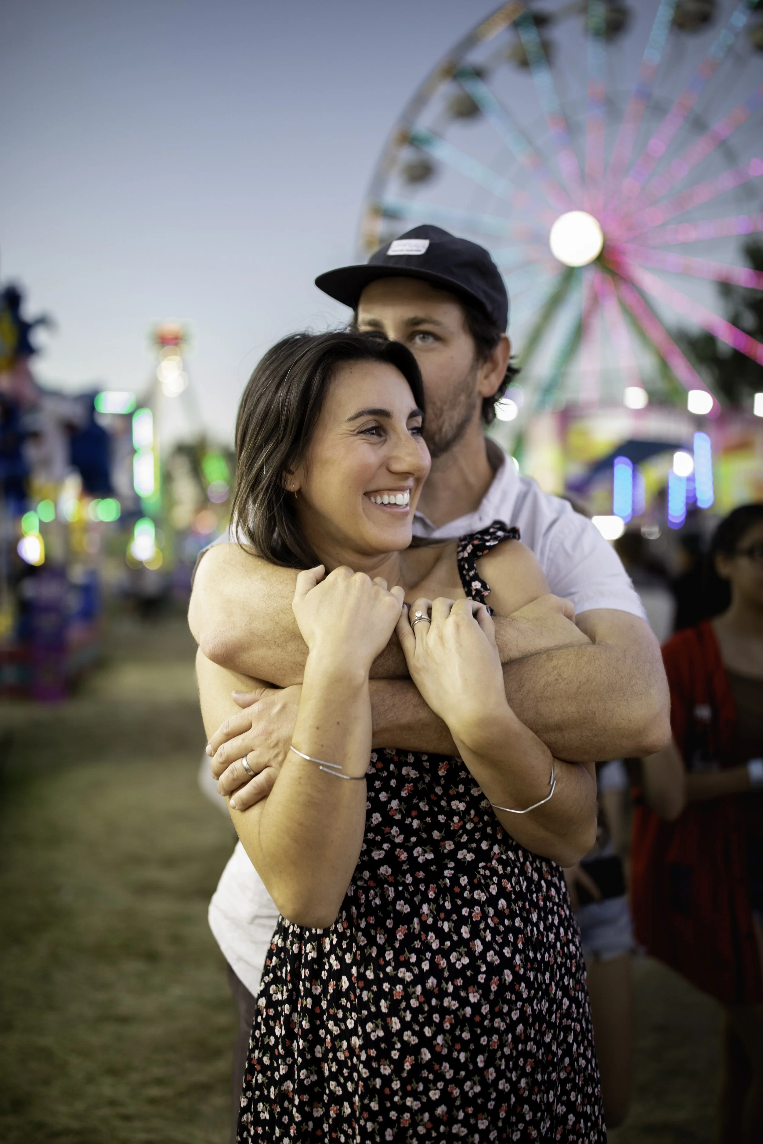A happy couple embracing at a fairground, with a Ferris wheel and amusement rides in the background during evening.