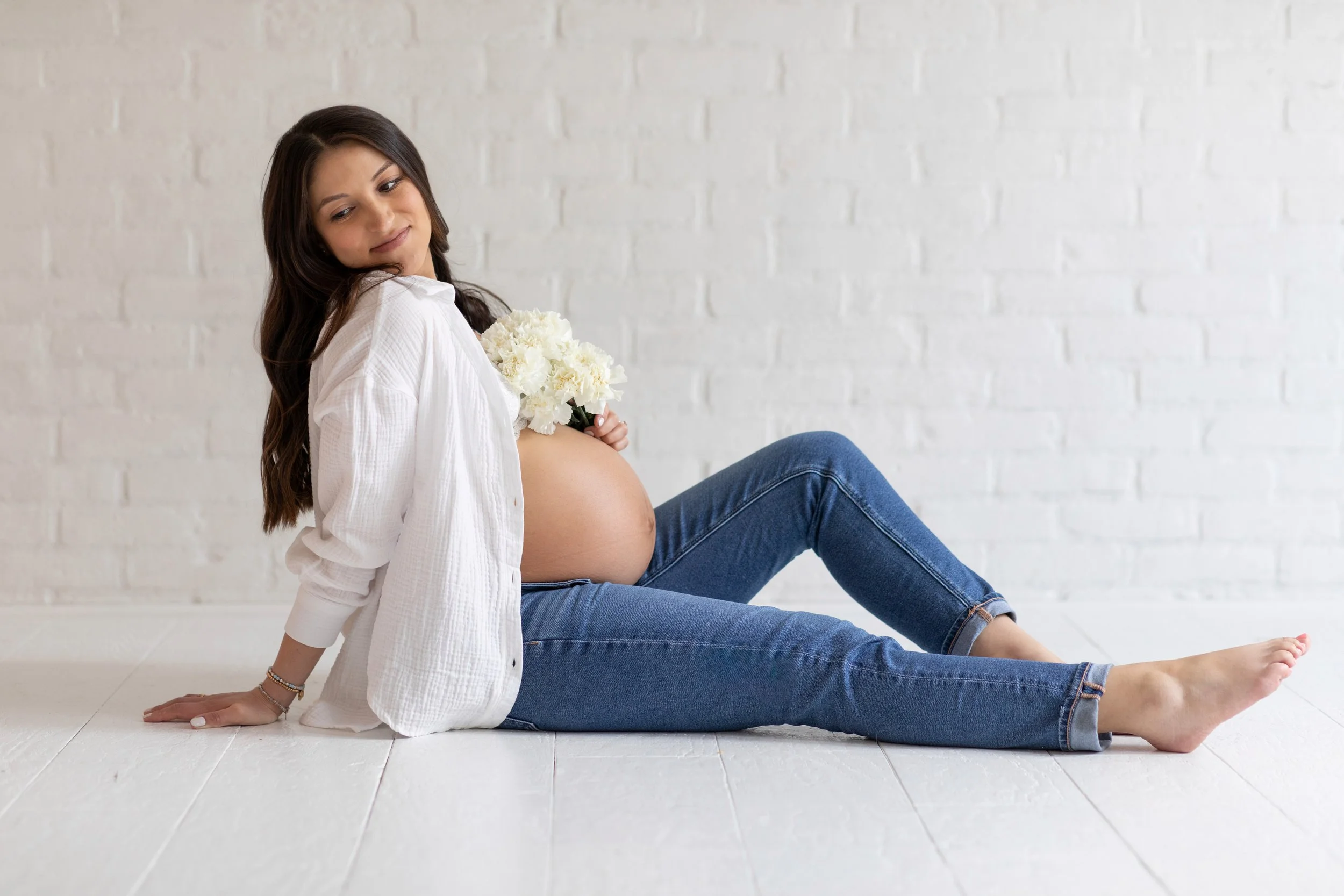 Pregnant woman sitting on the floor against a white brick wall, holding white flowers close to her pregnant belly.