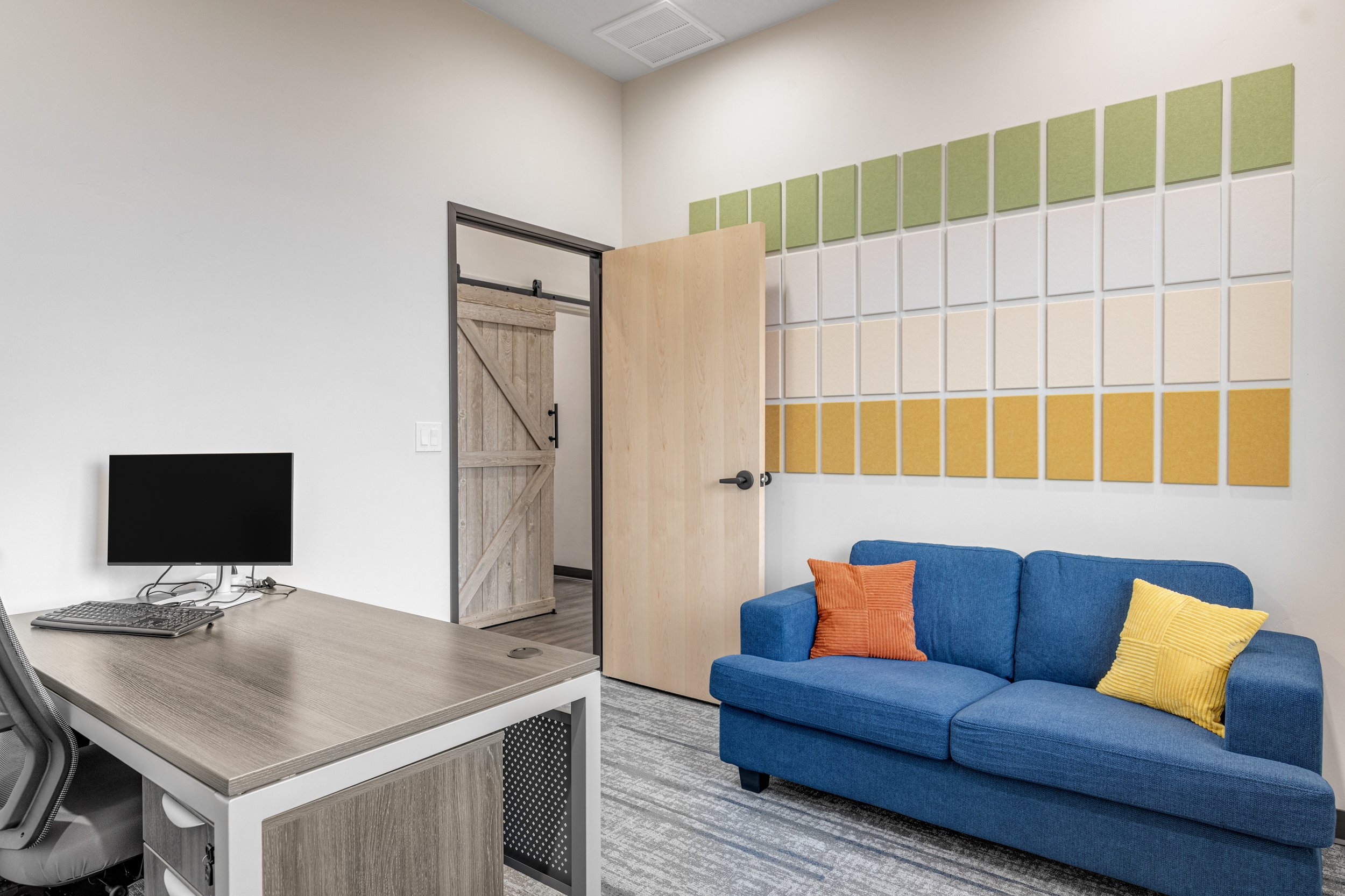 An office room with a work desk, a computer monitor, a gray office chair, a blue couch with orange and yellow pillows, a wooden sliding door, and colorful acoustic panels on the wall.