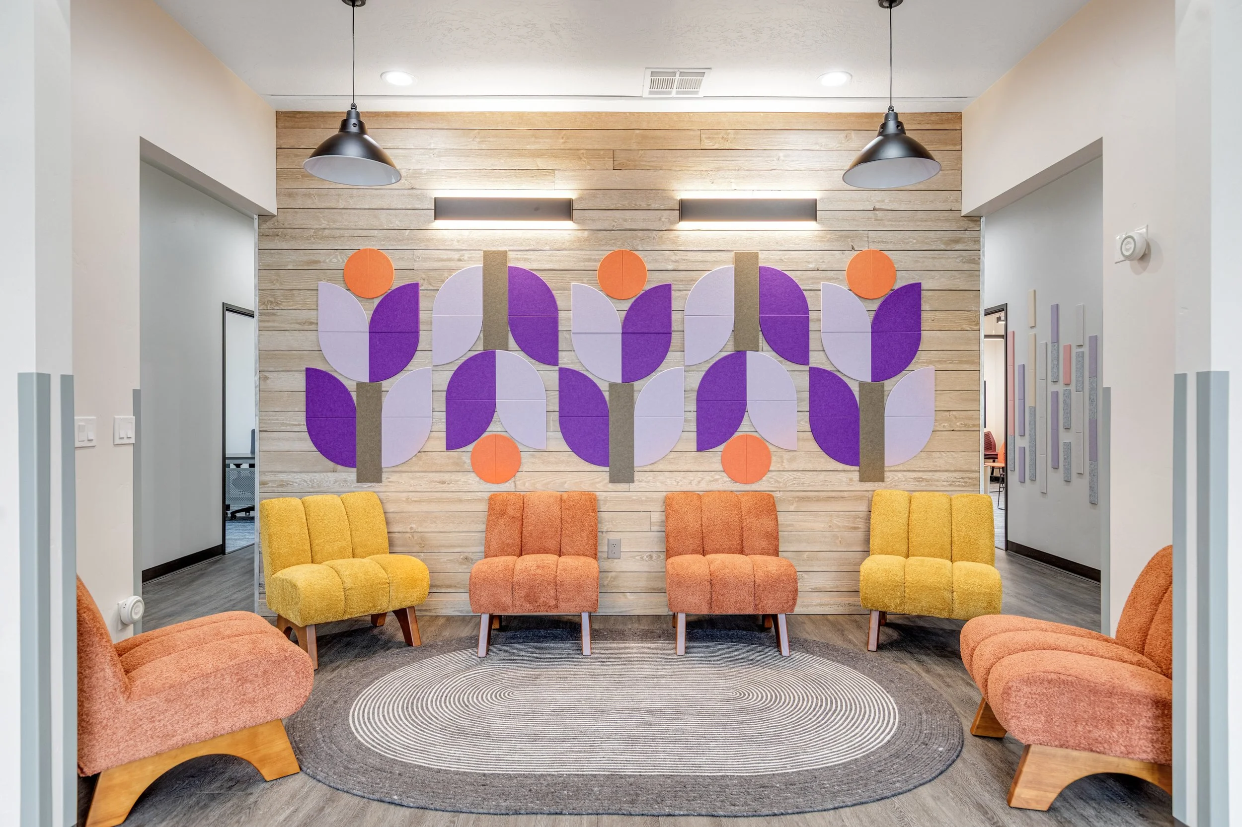 Reception or waiting area with colorful chairs, a circular rug, and a wooden wall with purple, lavender, and orange geometric art.