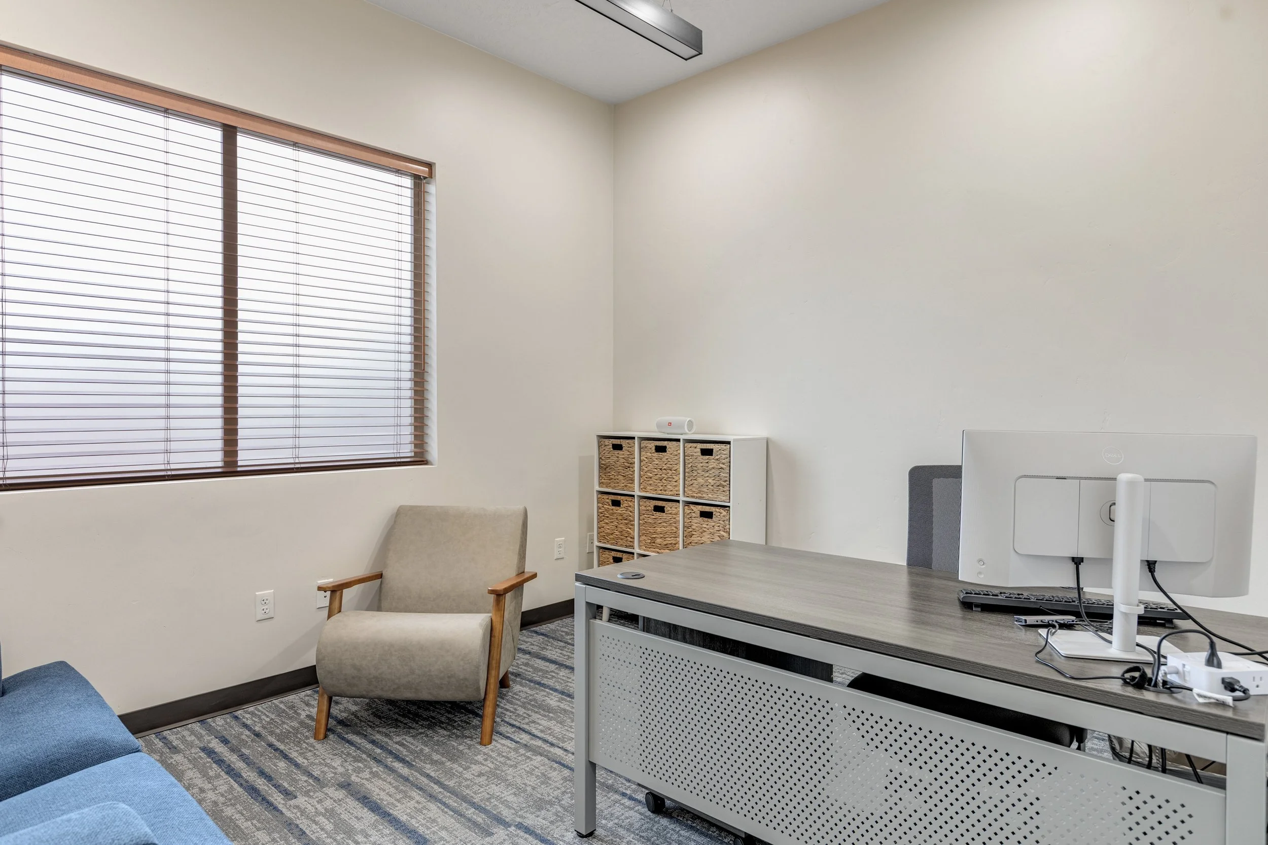 Empty modern office with a window, beige chair, blue sofa, white storage cabinet with wicker baskets, gray desk with a computer, and gray office chair.