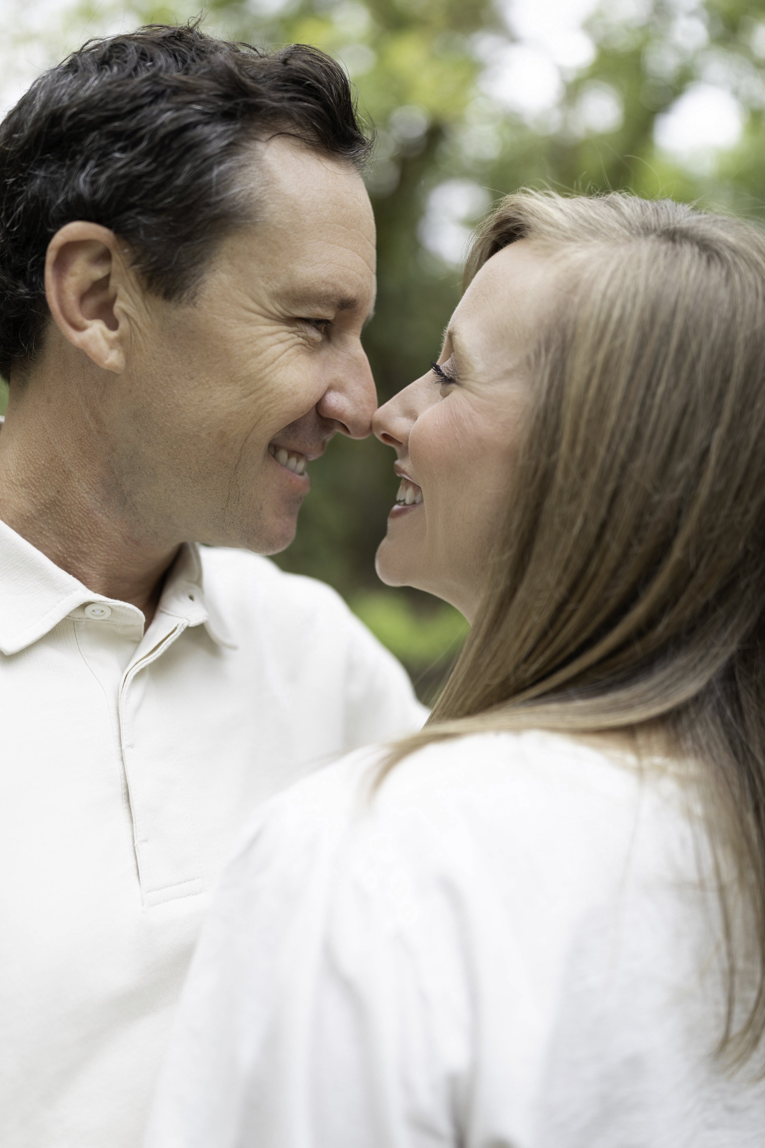 A couple is close together, touching noses and smiling at each other outdoors with blurred greenery in the background.