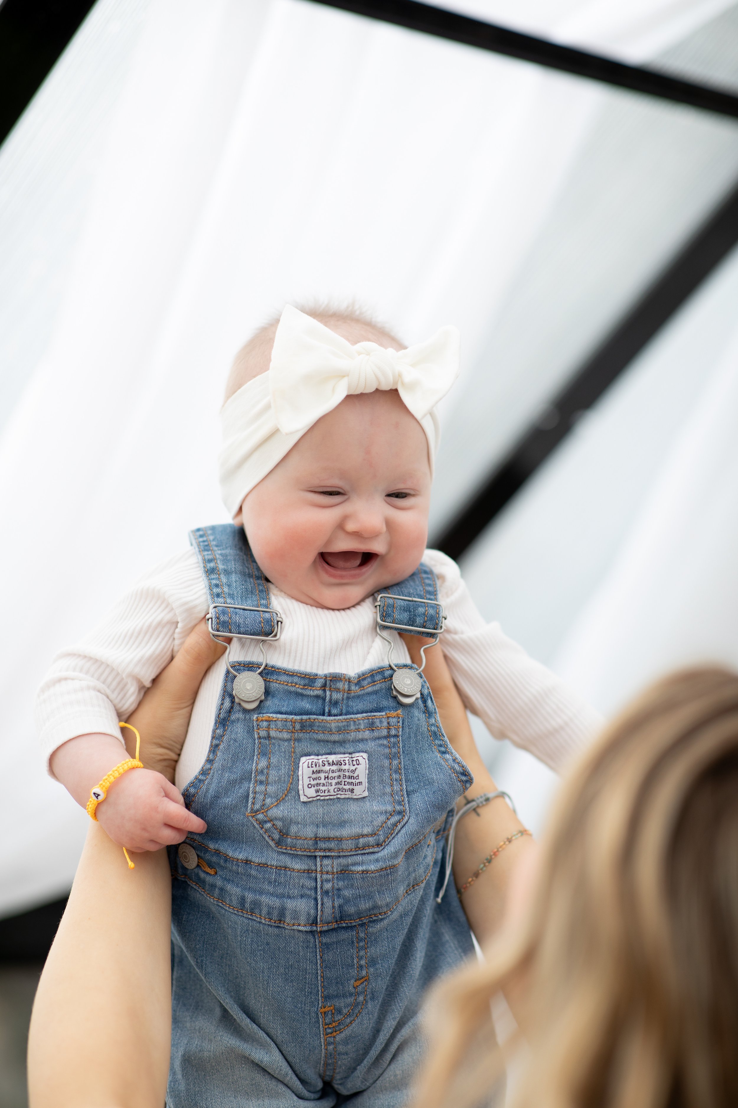 A smiling baby girl being lifted up, wearing a white headband with a bow, a white shirt, and denim overalls, inside a bright space with white and black structural elements.