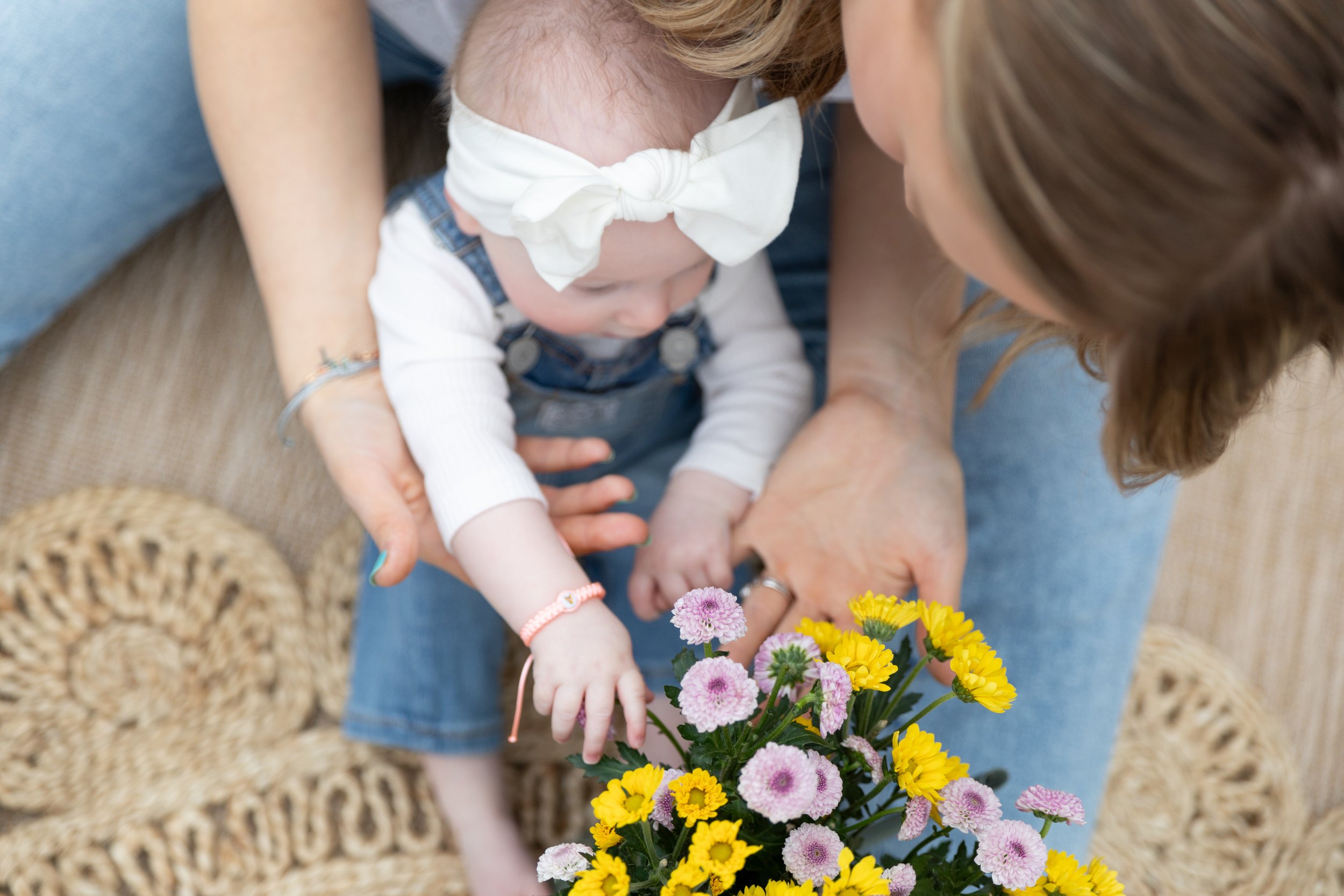 A woman and a young child, wearing denim overalls and a white top, are reaching out to touch a bouquet of yellow and pink flowers. The woman is guiding the child's hand gently.