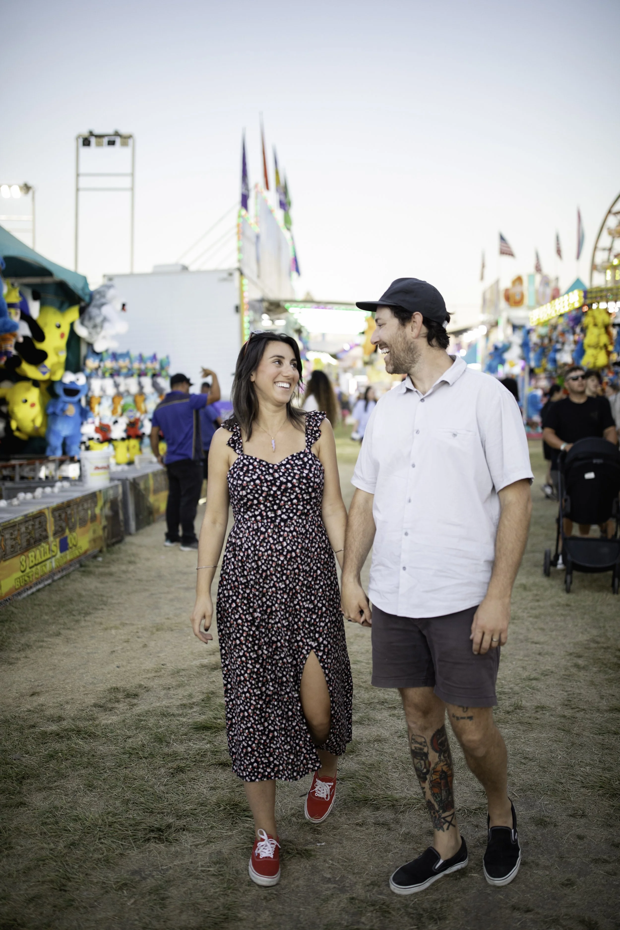 A couple walking hand-in-hand at a fairground with colorful game booths and carnival rides in the background, smiling at each other.