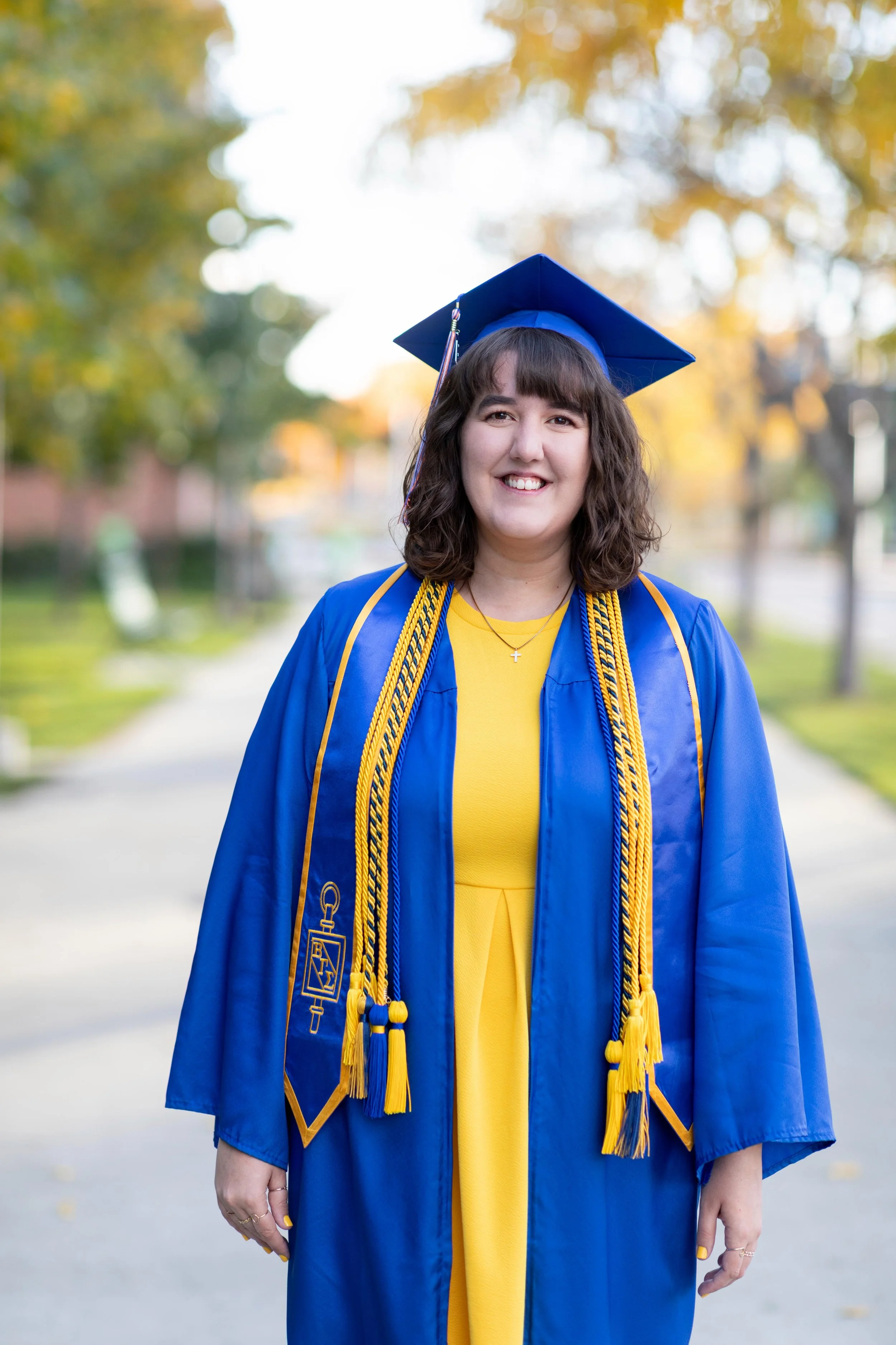 A young woman in a yellow dress and blue graduation gown and cap, standing outdoors, smiling.