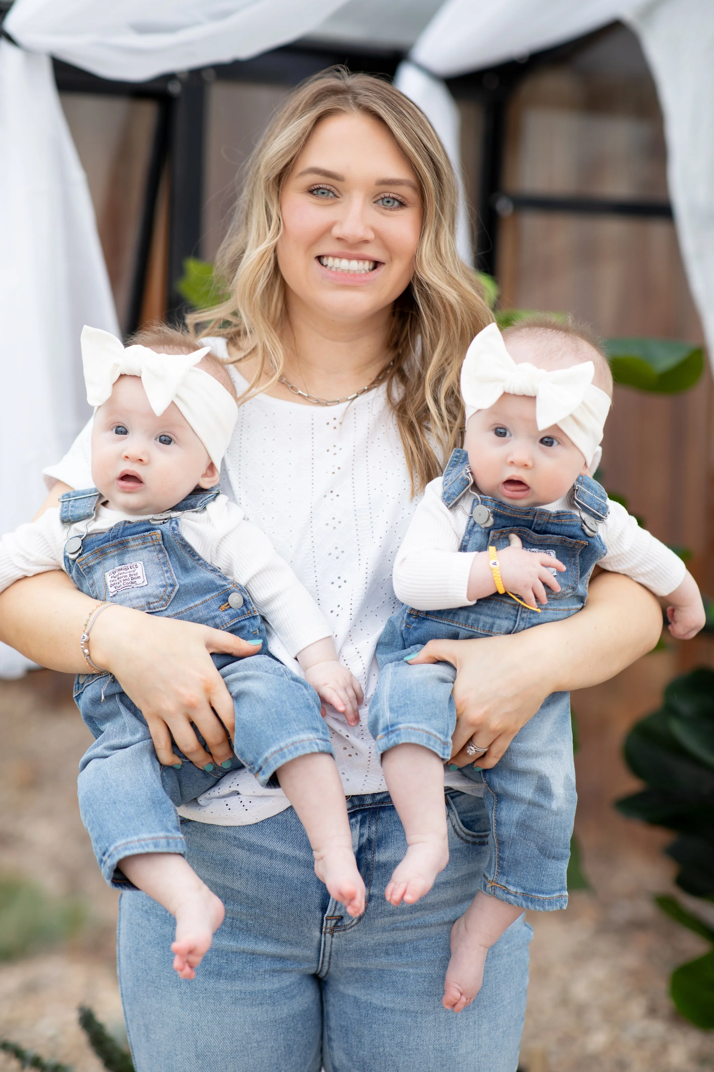 A smiling woman holding two babies dressed in denim overalls with white bows in their hair, standing outdoors with greenery and a white tent in the background.