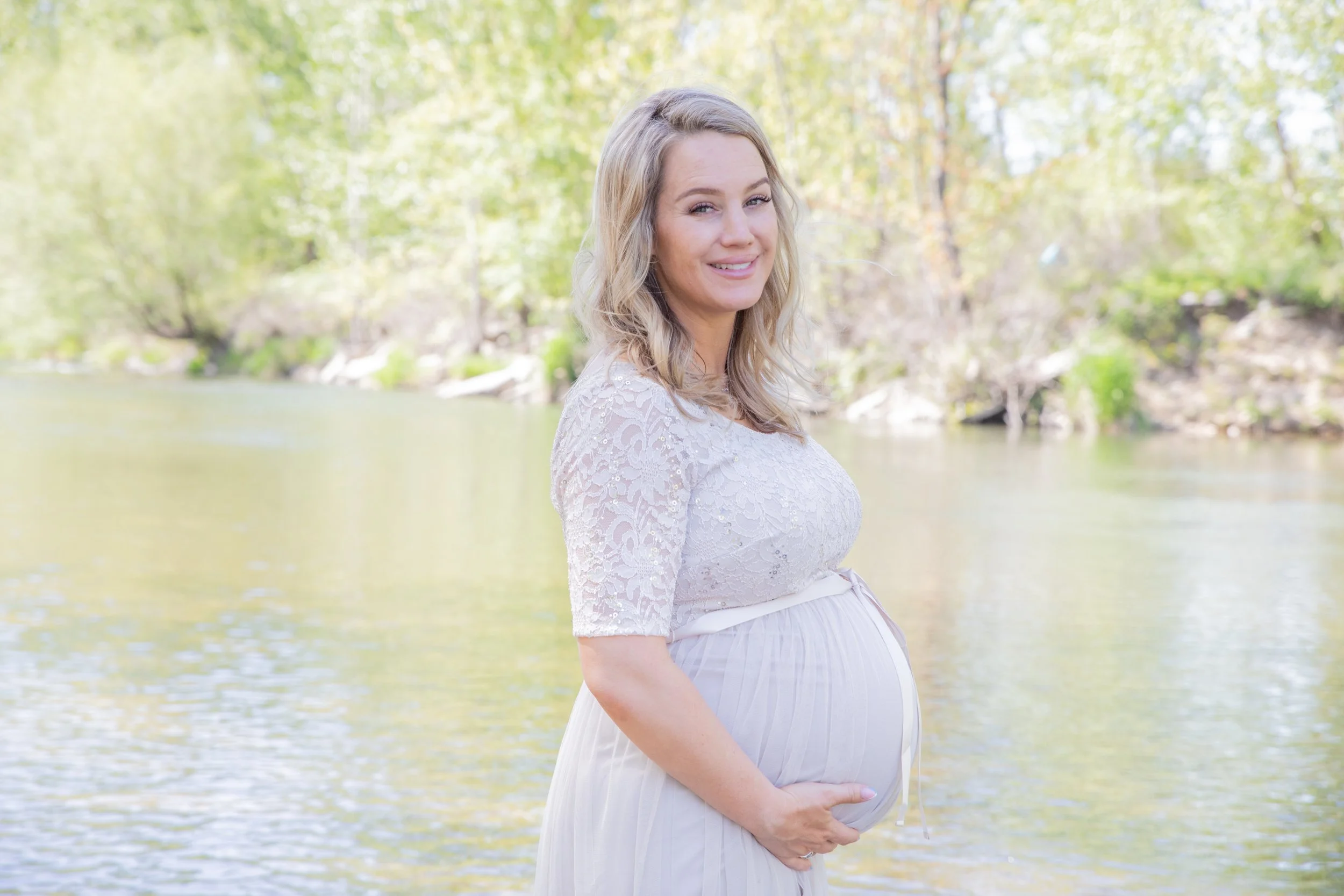 A pregnant woman standing by a river, smiling and holding her belly, surrounded by trees and greenery.