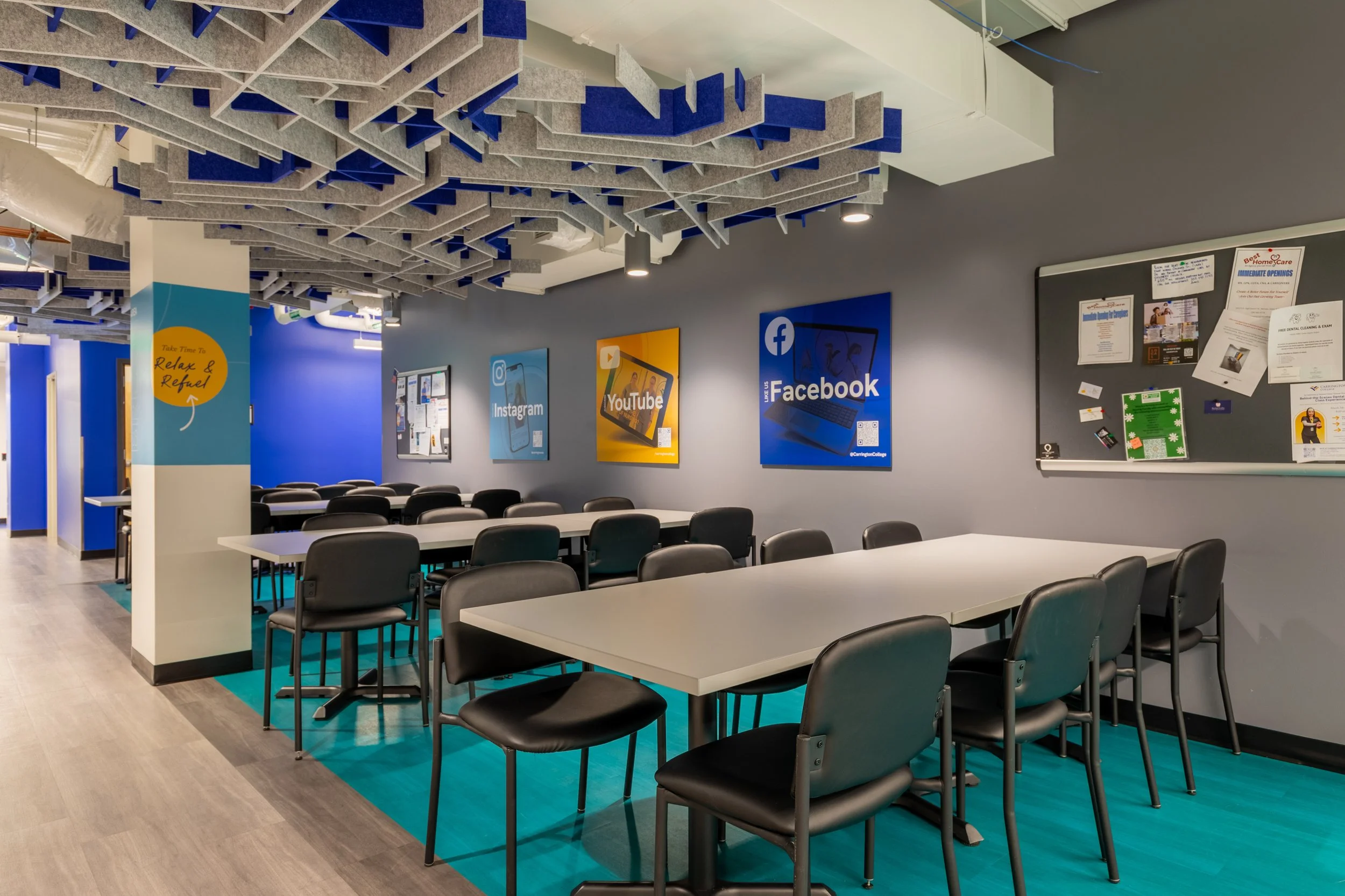An interior classroom or meeting room with several tables and chairs. The walls display posters of social media platforms like Instagram, YouTube, and Facebook. There’s a bulletin board with notices and papers, and the ceiling is decorated with acous