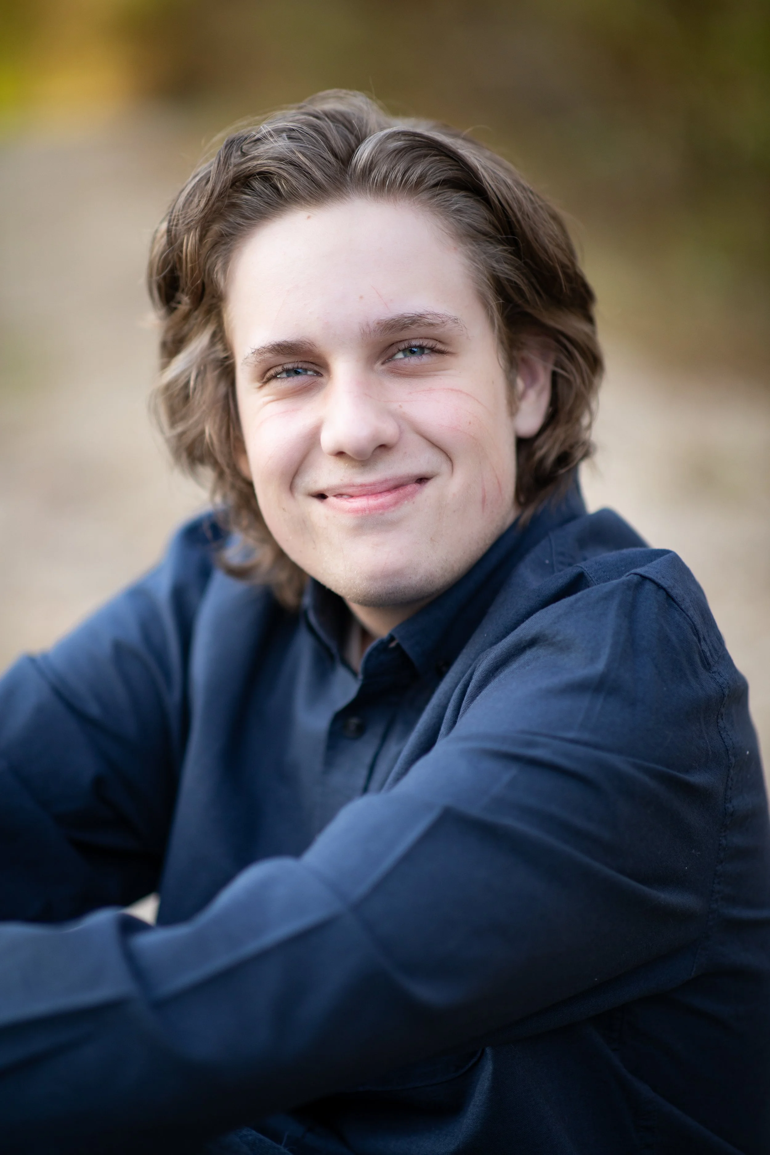 Close-up portrait of a young man with shoulder-length brown wavy hair, smiling gently outdoors in a natural setting with blurred background.