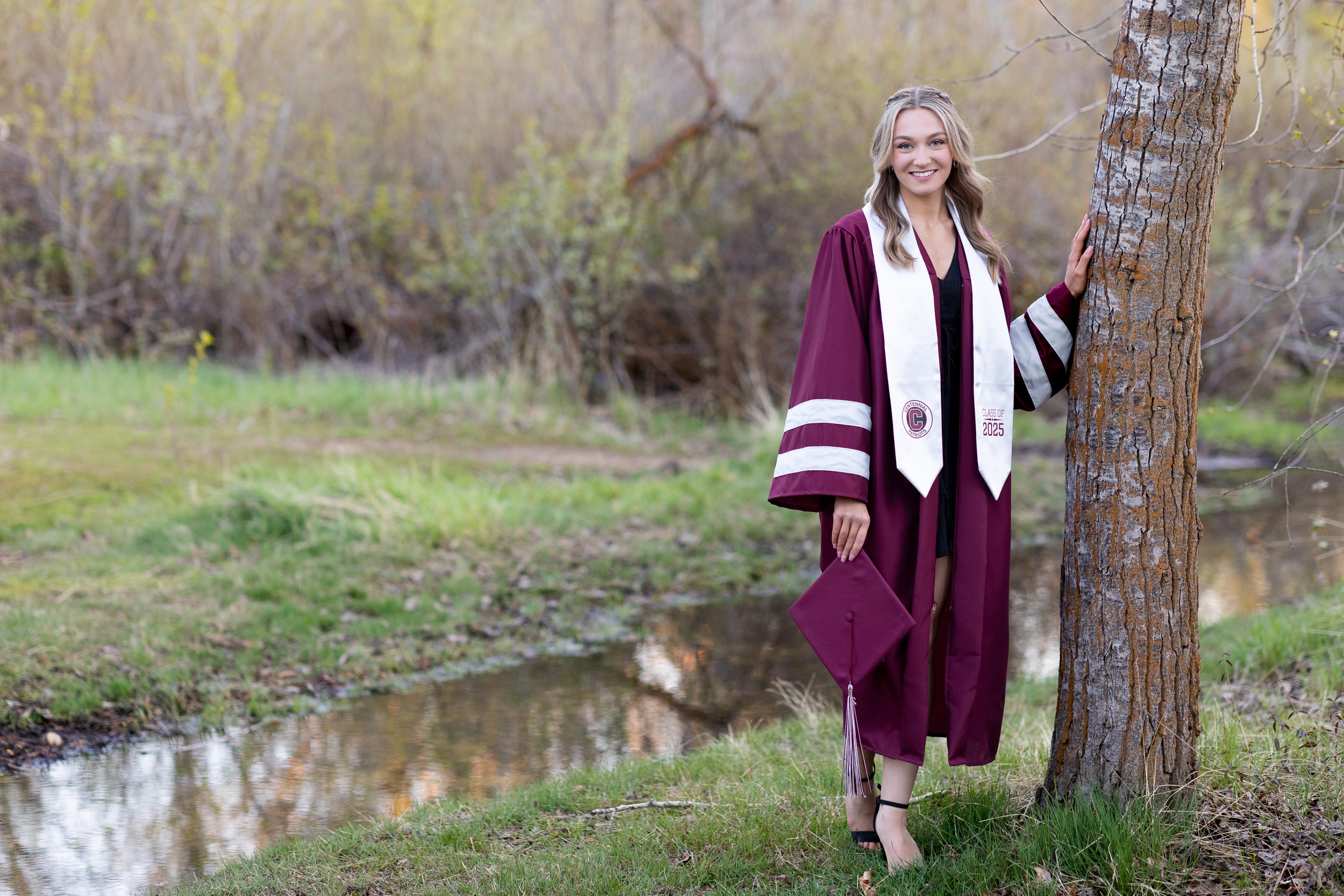 A young woman in a maroon and white graduation gown holding a mortarboard cap stands outdoors near a tree and a small creek, smiling.