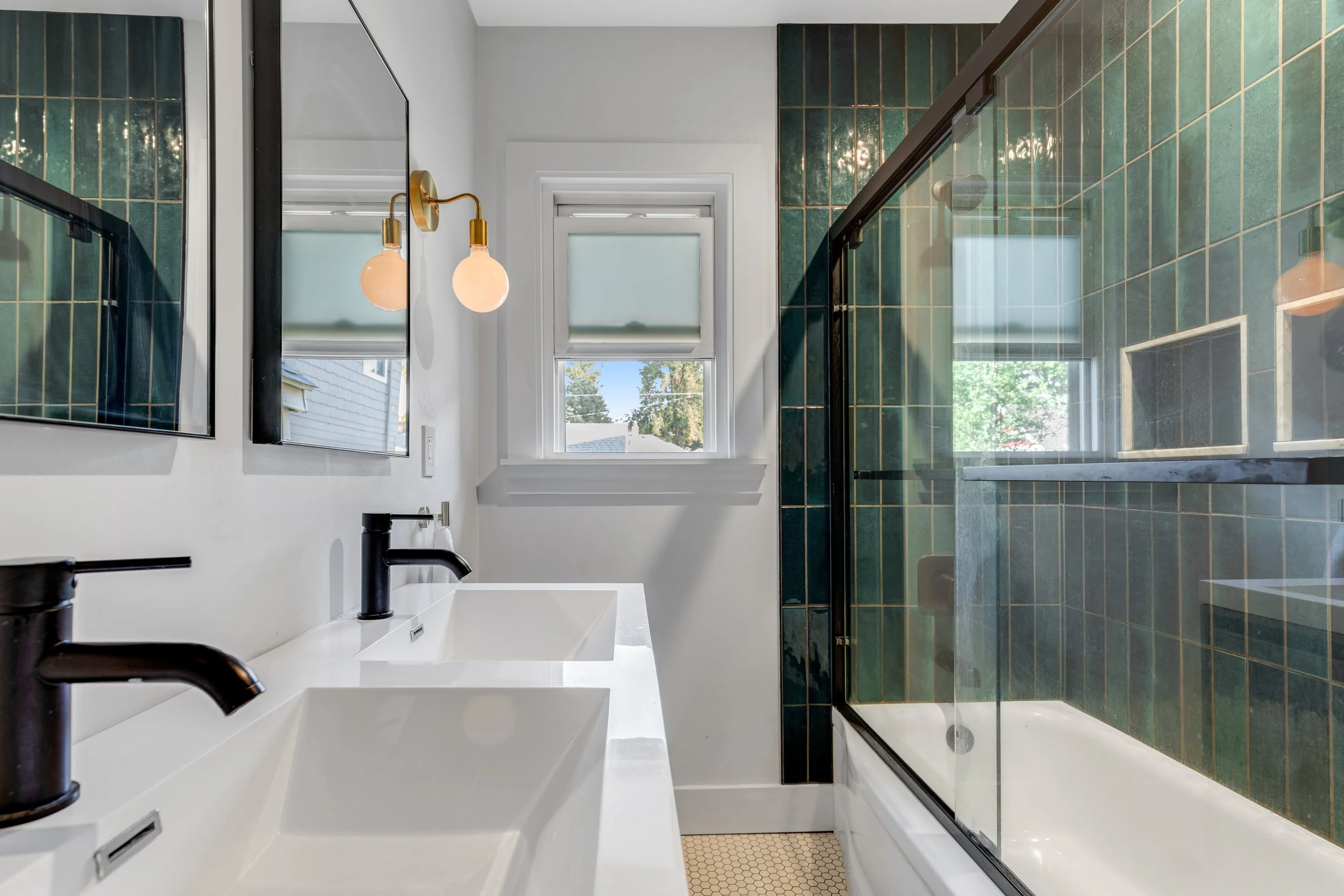 Bathroom with double white sinks, black faucets, a mirror, green tiled shower with glass door, small window with blinds, and white walls.