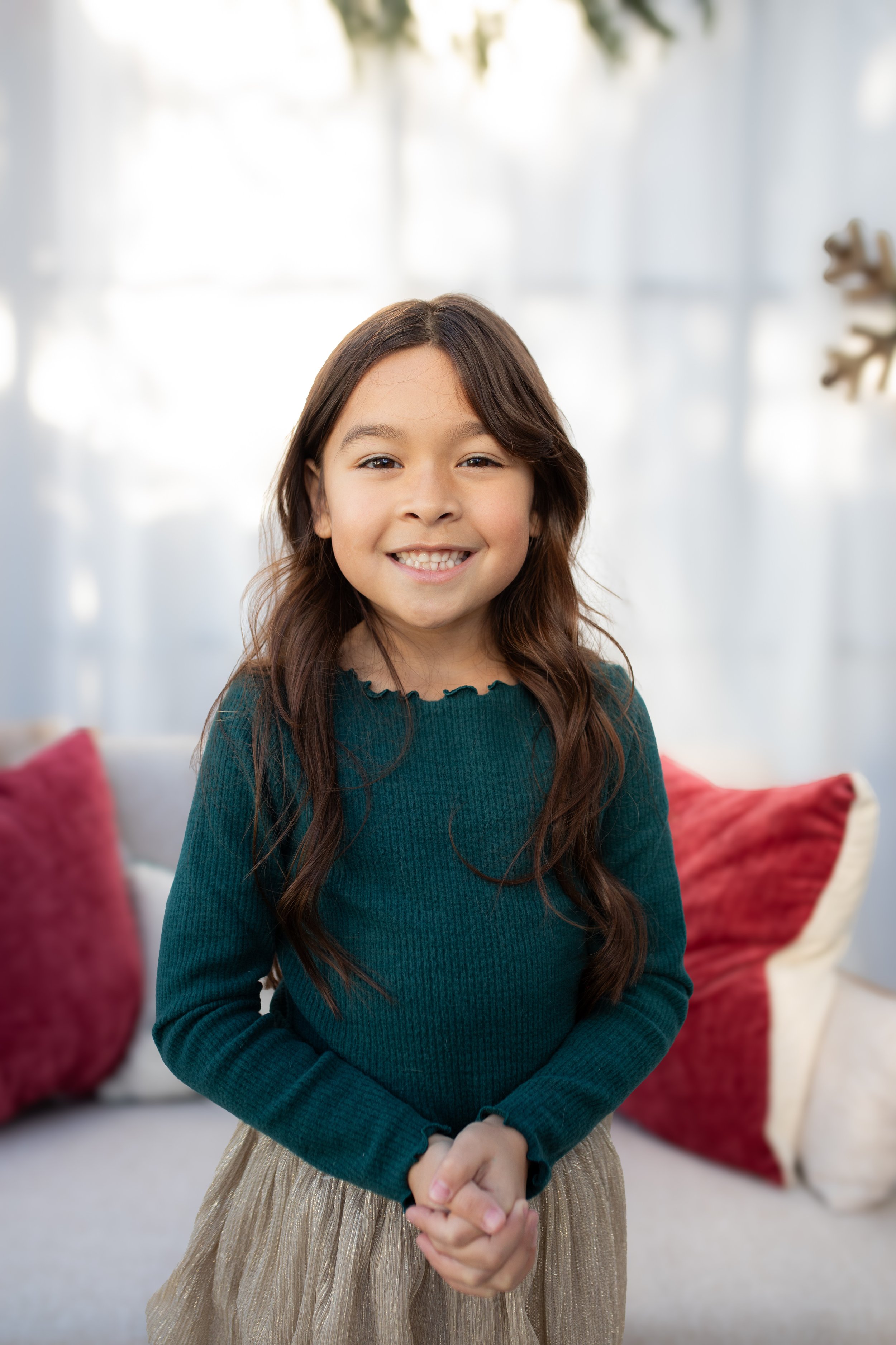 A smiling young girl with long brown hair, wearing a green sweater and a beige skirt, standing in a bright living room with red cushions on a white sofa and a Christmas-themed decoration in the background.