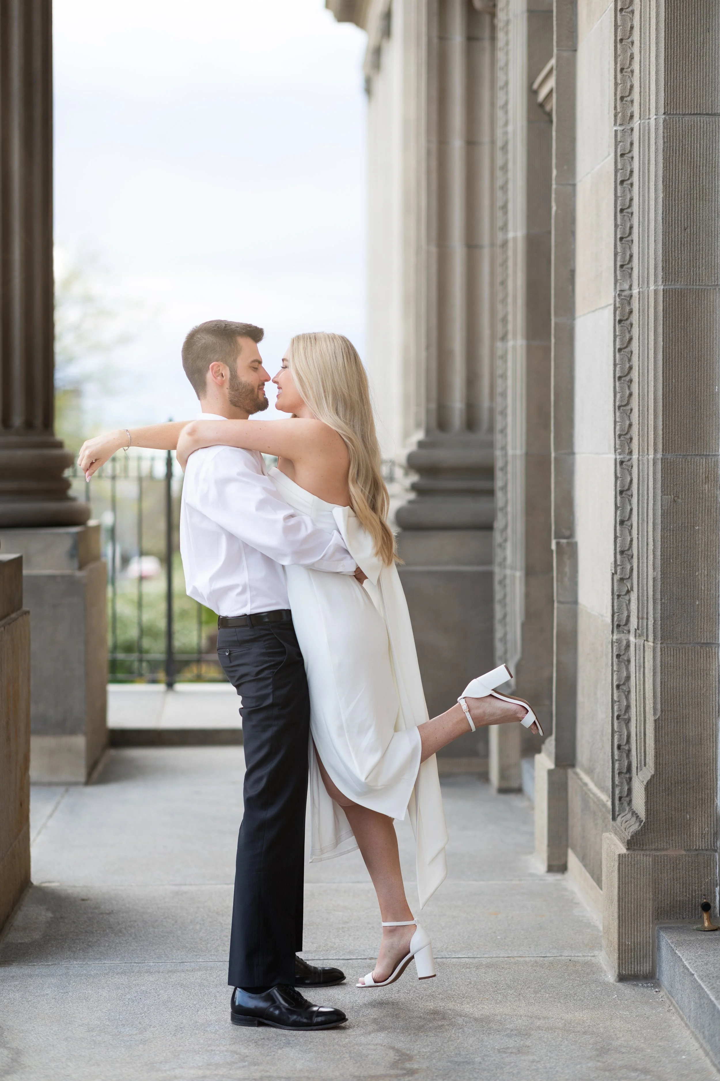 A couple dressed in formal white and black clothes, outside near stone columns, sharing a romantic moment. The woman is being lifted with her arms around the man's neck and one leg bent behind her, smiling and looking at each other.