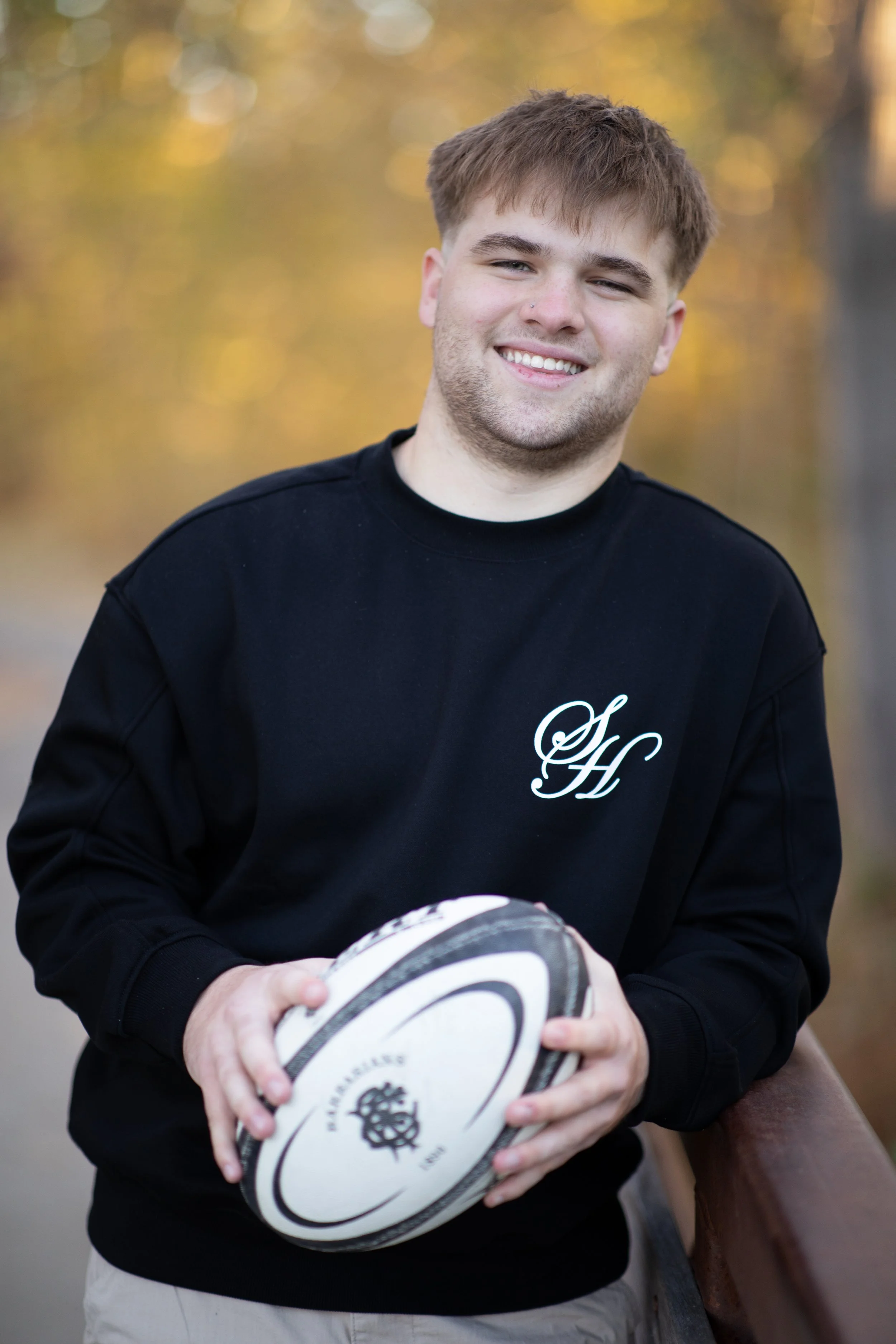 A young man with light brown hair, fair skin, and a slight beard, smiling and holding a rugby ball outdoors during fall, standing on a bridge with autumn trees in the background.