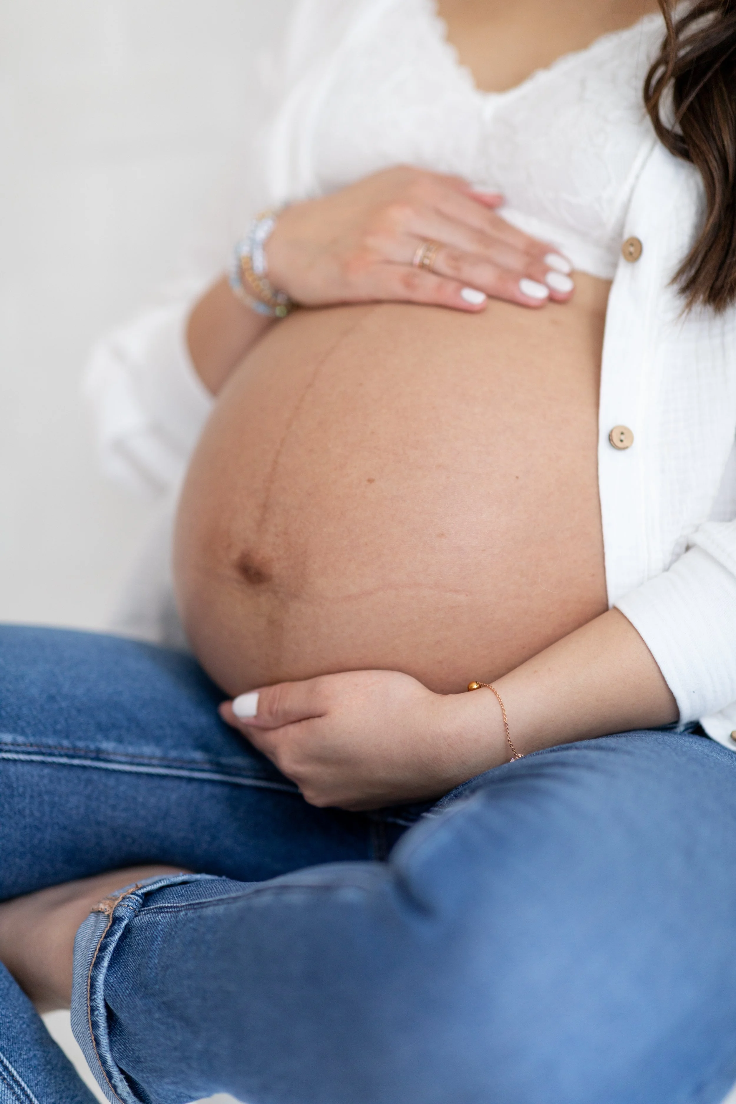 A pregnant woman is sitting cross-legged, holding her belly with both hands. She is wearing a white shirt and blue jeans, with her left hand resting on top of her belly and her right hand supporting underneath. The background is neutral and out of fo