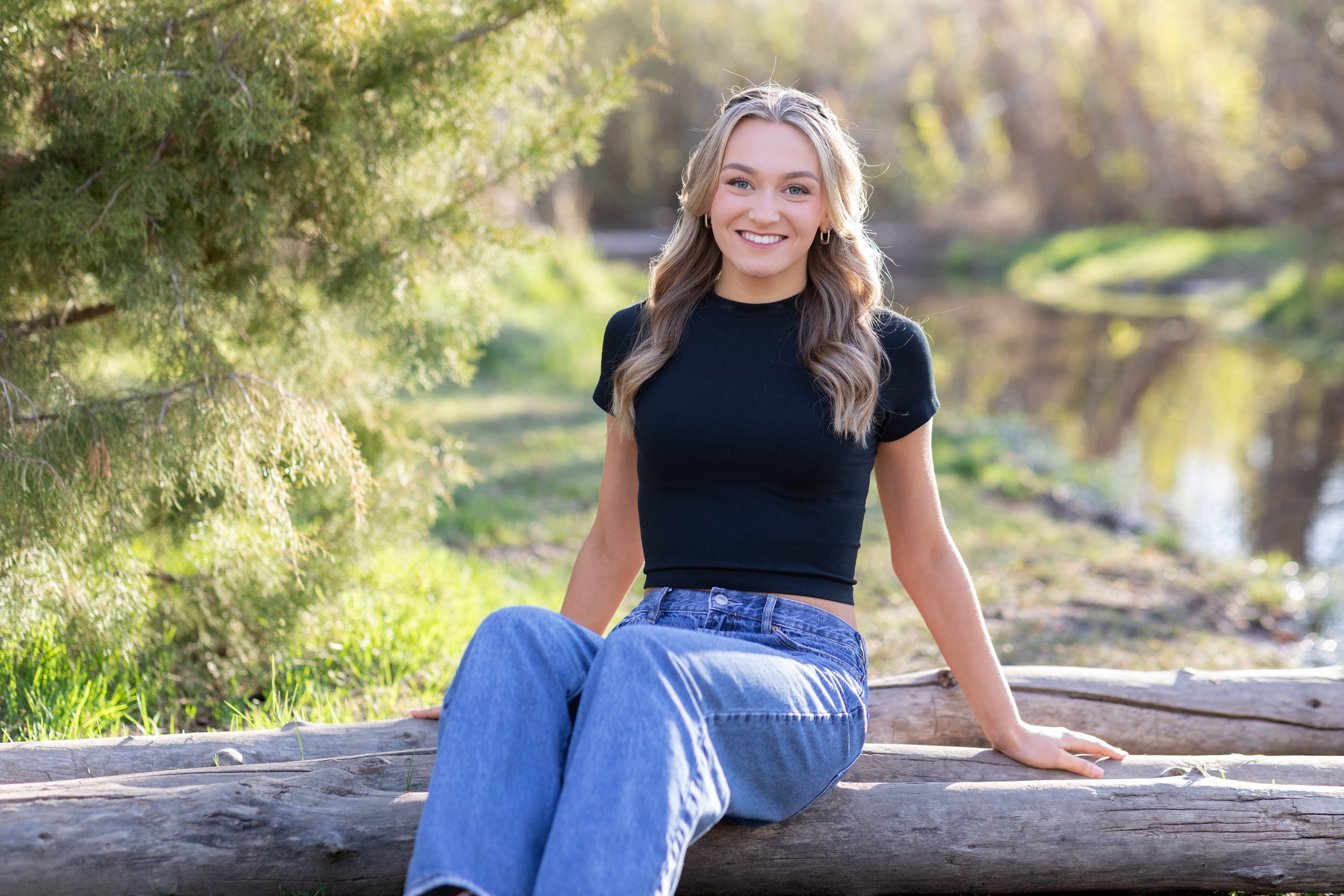 A young woman with long wavy hair, wearing a black short-sleeve top and blue jeans, sitting on a log in a natural outdoor setting with green trees and a small body of water in the background, smiling at the camera.