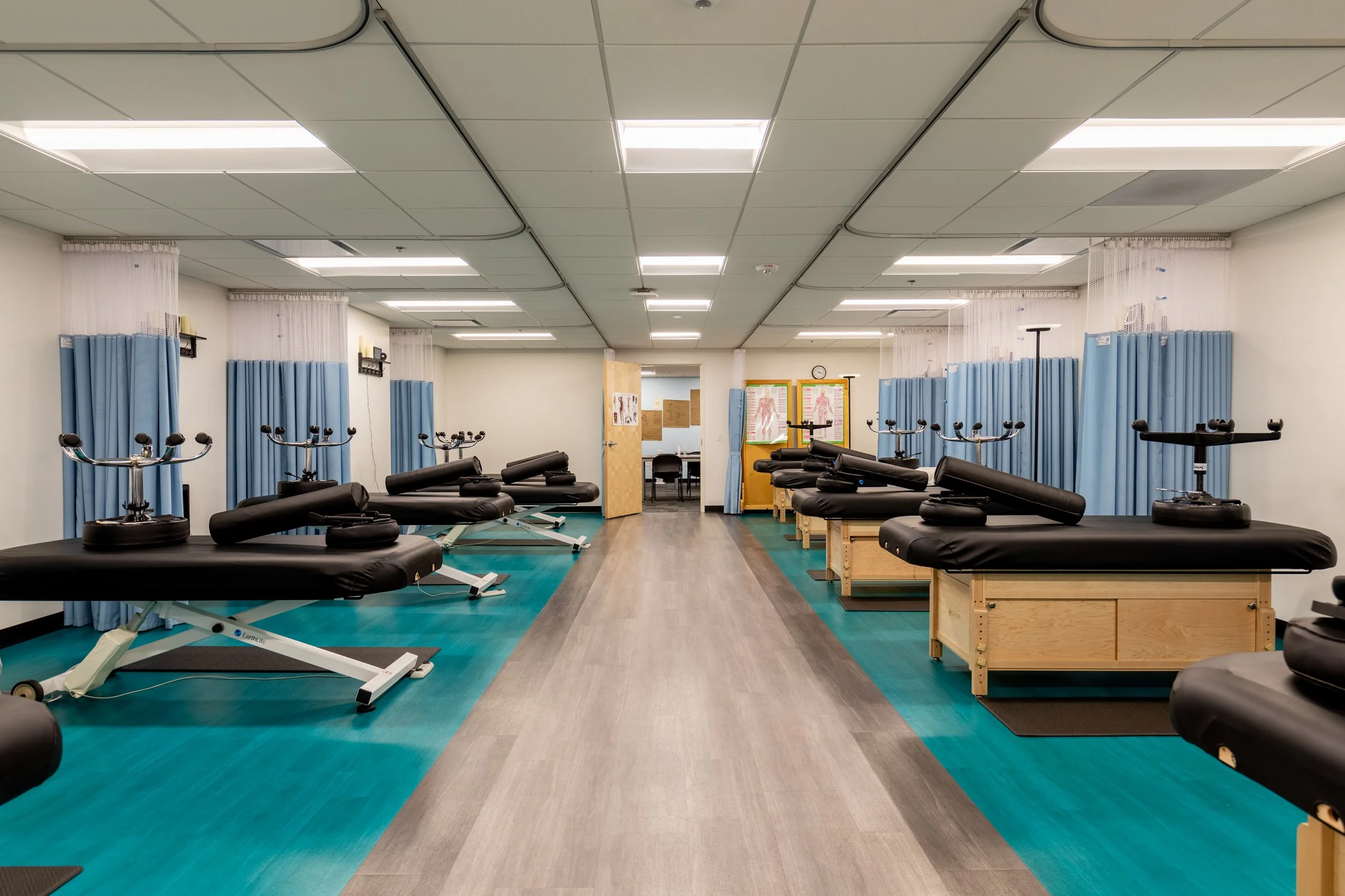 Empty physical therapy or rehabilitation treatment room with adjustable massage tables, blue privacy curtains, and medical charts on the walls.