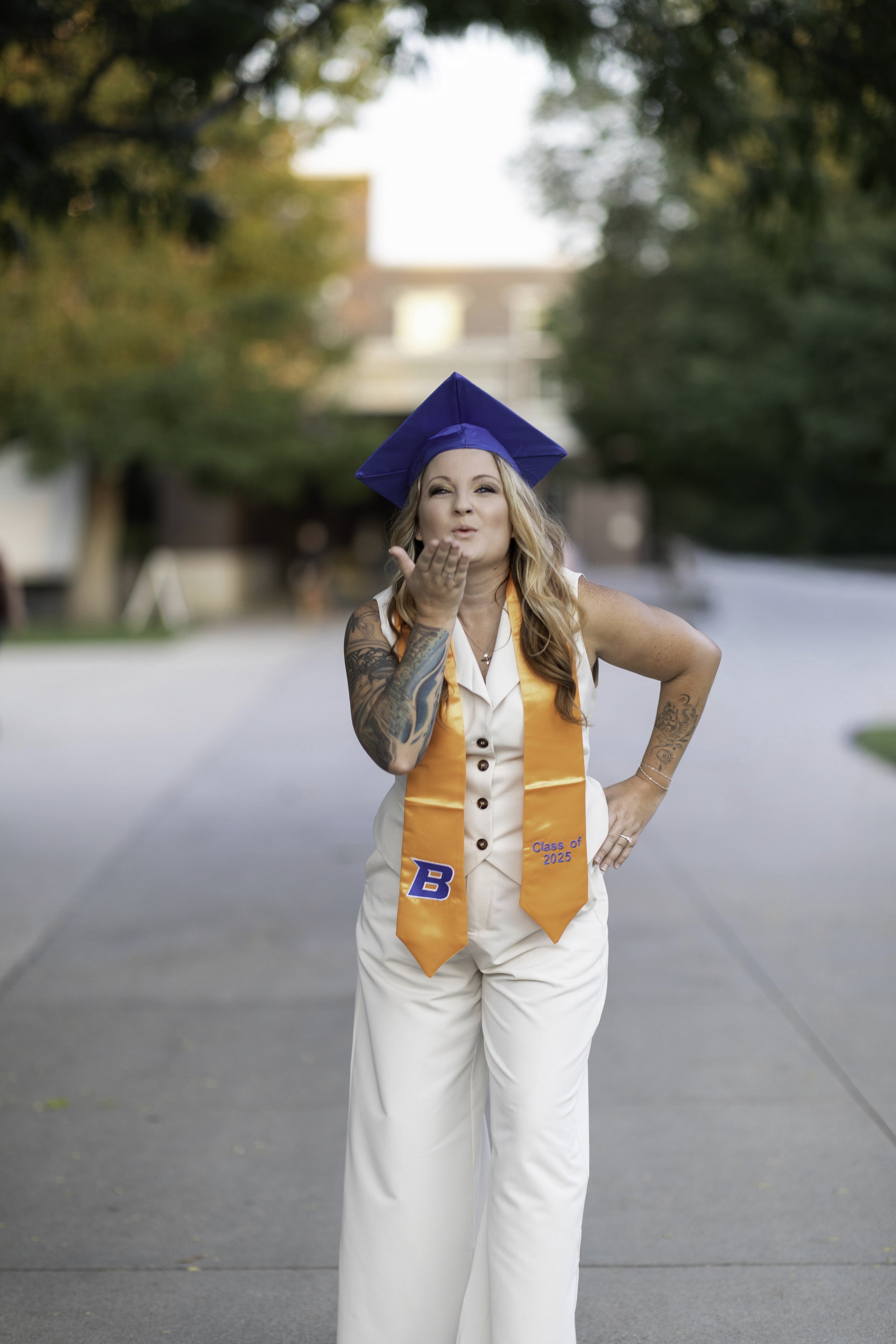 Woman in a cream-colored jumpsuit and blue graduation cap blowing a kiss, wearing an orange graduation stole that says 'Class of 2025' and has a blue 'B' on it, standing outdoors on a sidewalk.