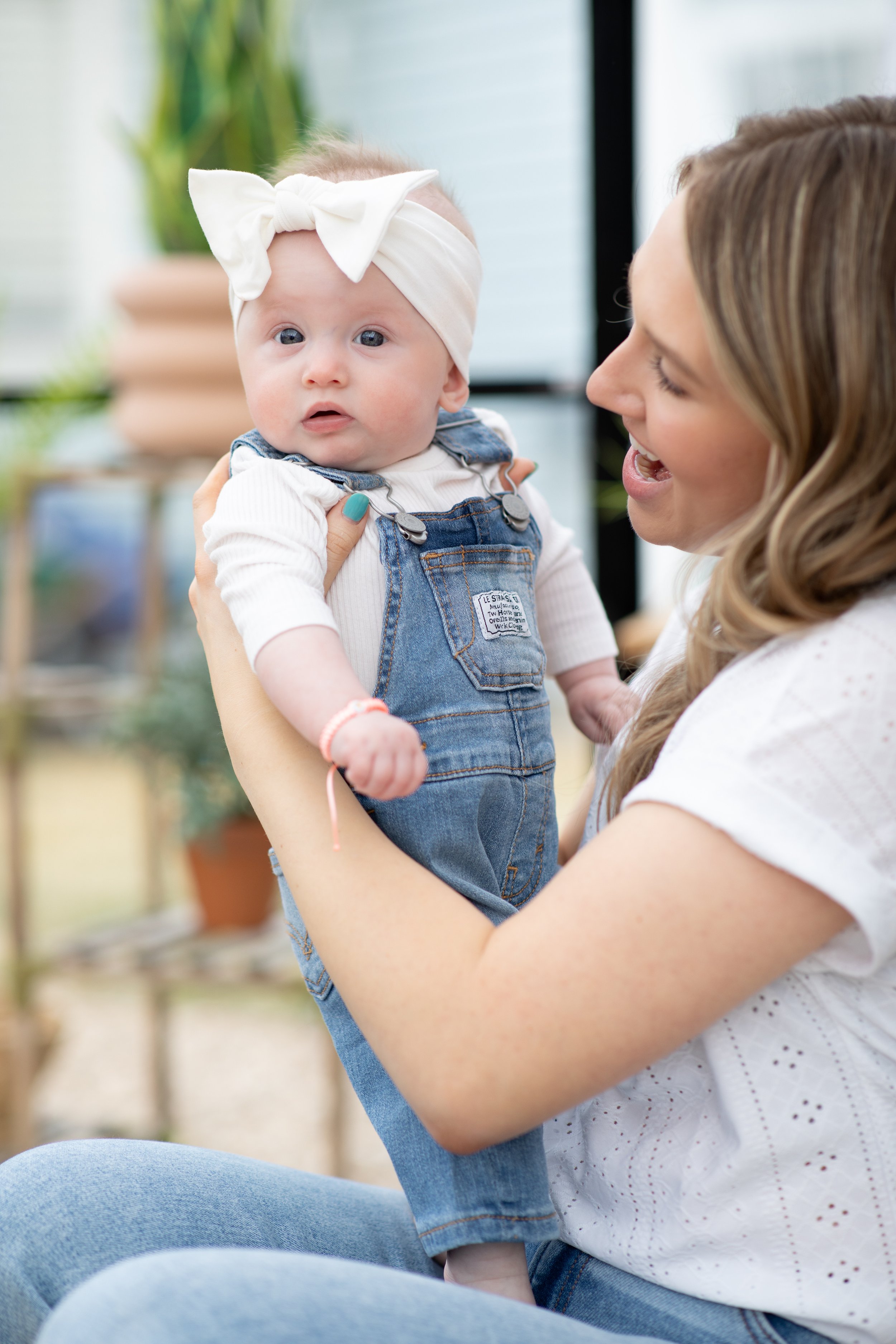 A woman holding a baby girl with a white headband and denim overalls in a bright indoor space.