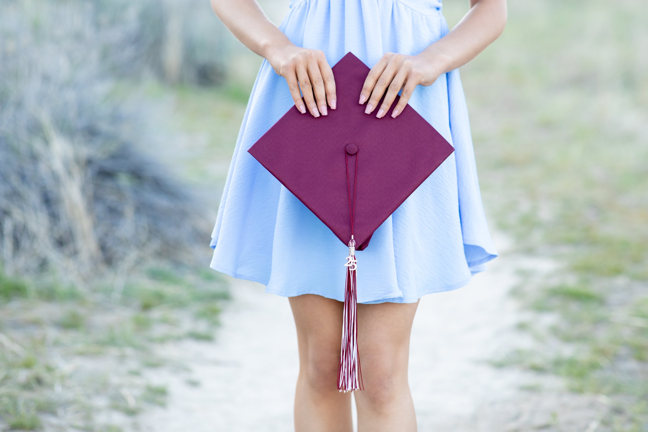 Person holding a maroon graduation cap with a tassel, standing outdoors on a grassy path.
